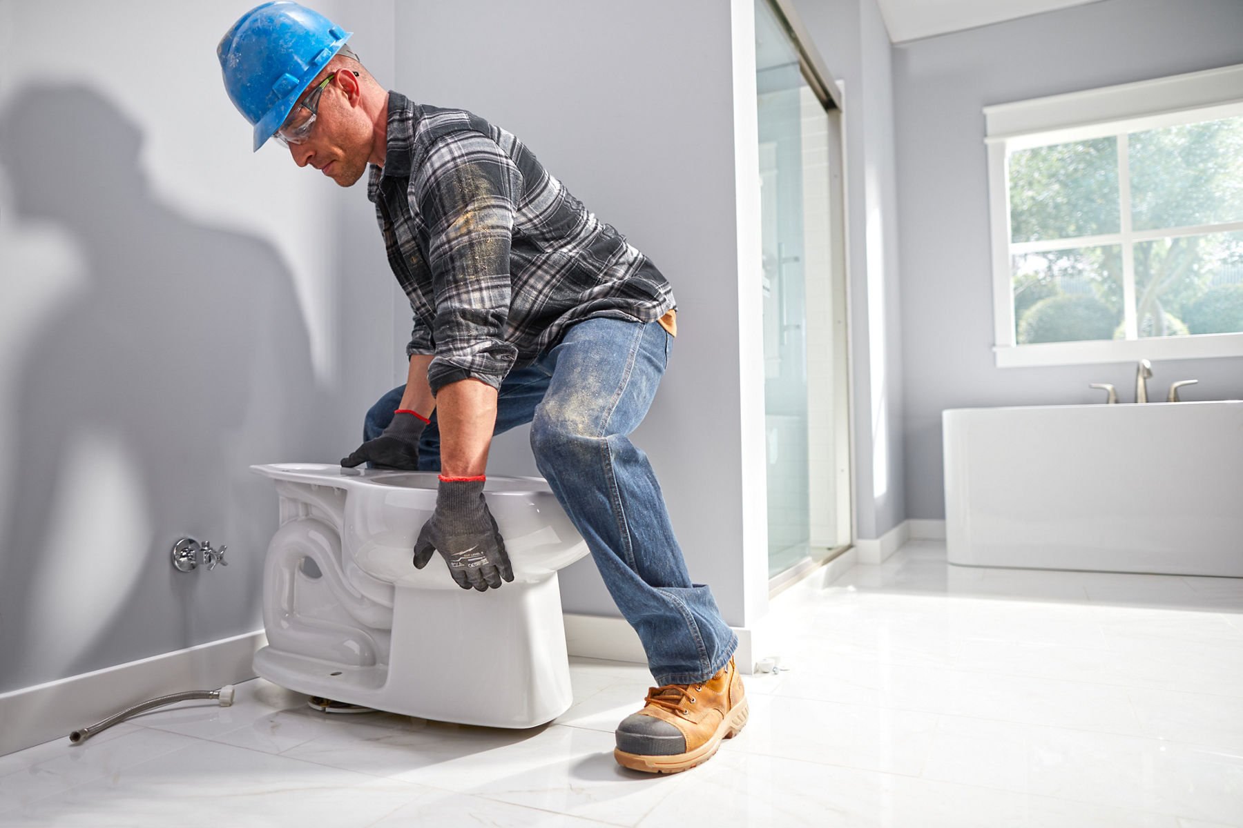 A man in a hardhat and yellow safety vest installing a white toilet bowl base in a pale grey bathroom.