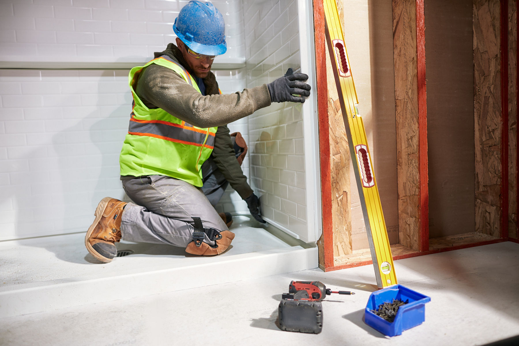 A man in a hardhat and yellow safety vest installing a white shower panel in a roughed-out bathroom.