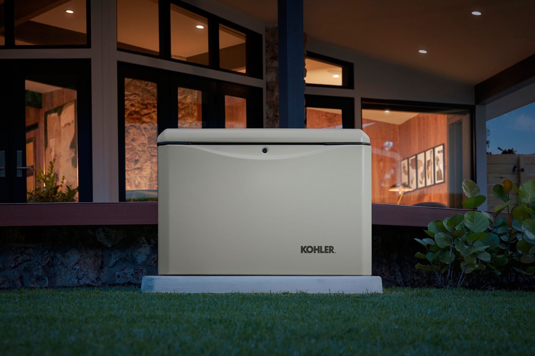 A tan colored home generator outside a home at night with its windows illuminated.