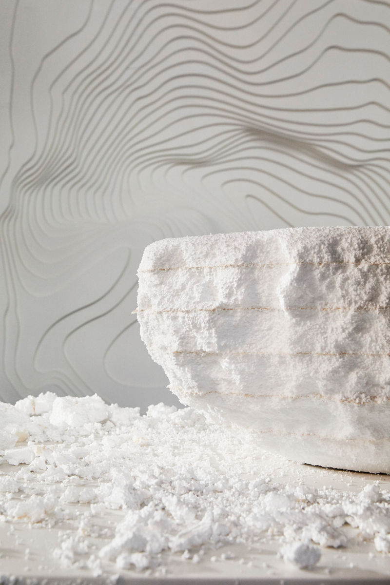 A textured white bowl-like sculpture sits on a surface scattered with white granular material, against a backdrop featuring a wavy, embossed pattern on a light-colored wall.