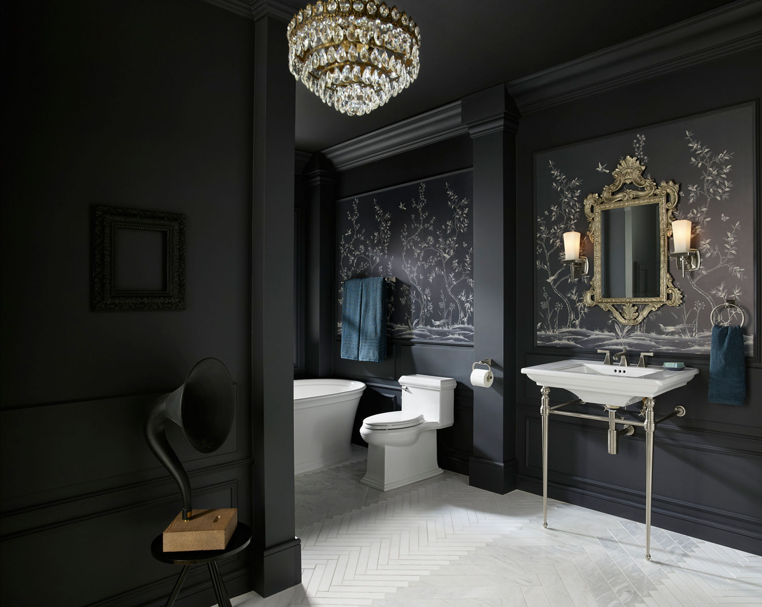 A Memoirs console table sink, toilet, and freestanding bath in a bathroom with dark gray walls with ornate white plant life drawings and an ornate gold-framed mirror.