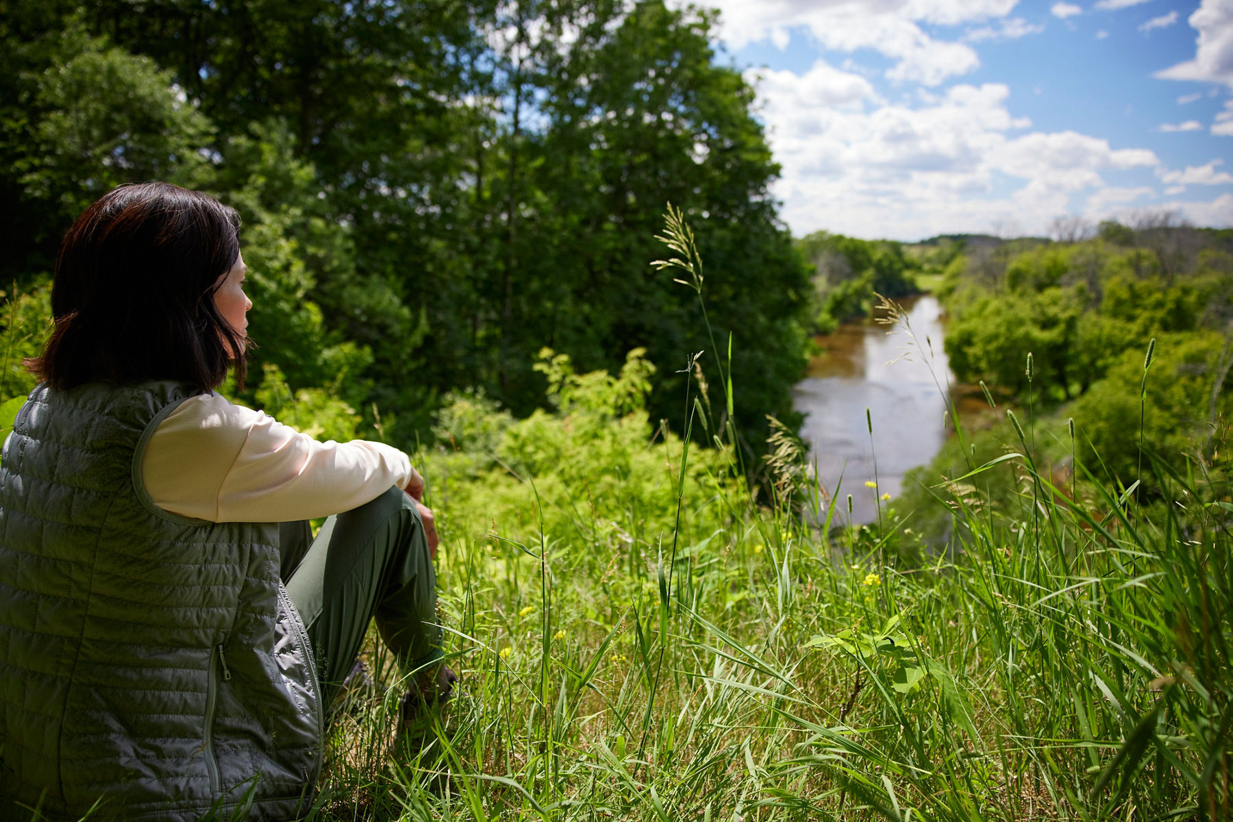 Image of woman sitting in natural tall grass along the edge of a river, looking down at the water and trees lining the river banks.
