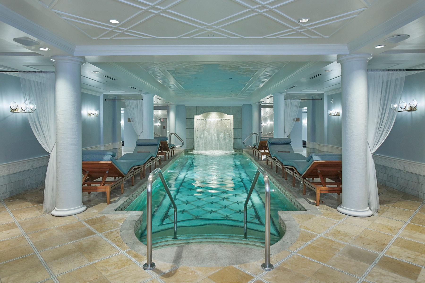 Wide image of the soaking pool inside the Kohler Waters Spa, showing crystal clear water with a patterned tile bottom of the pool, and white pillars and white draped linens around the sitting areas of the pool.