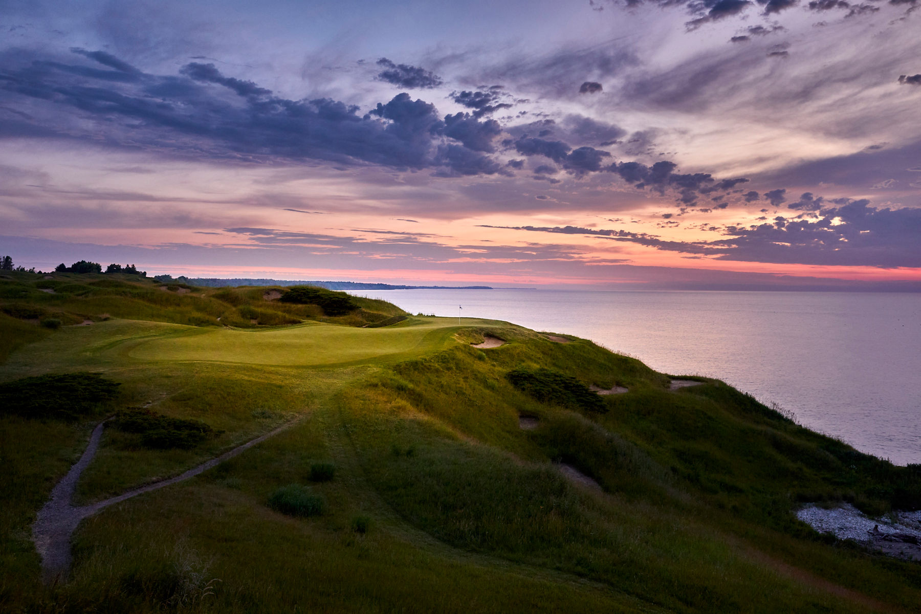  Wide landscape image of the green at a Kohler golf course, showing the sunset skies and clouds across the lake in the distance.