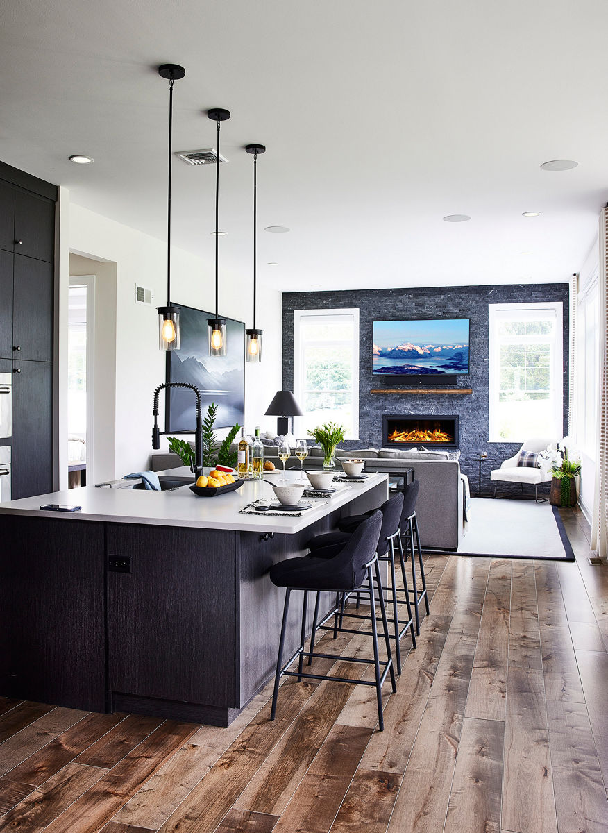 A large room comprising a kitchen island with an integrated faucet and sink and seating in the foreground and a living area with a sofa, fireplace and large windows in the background.