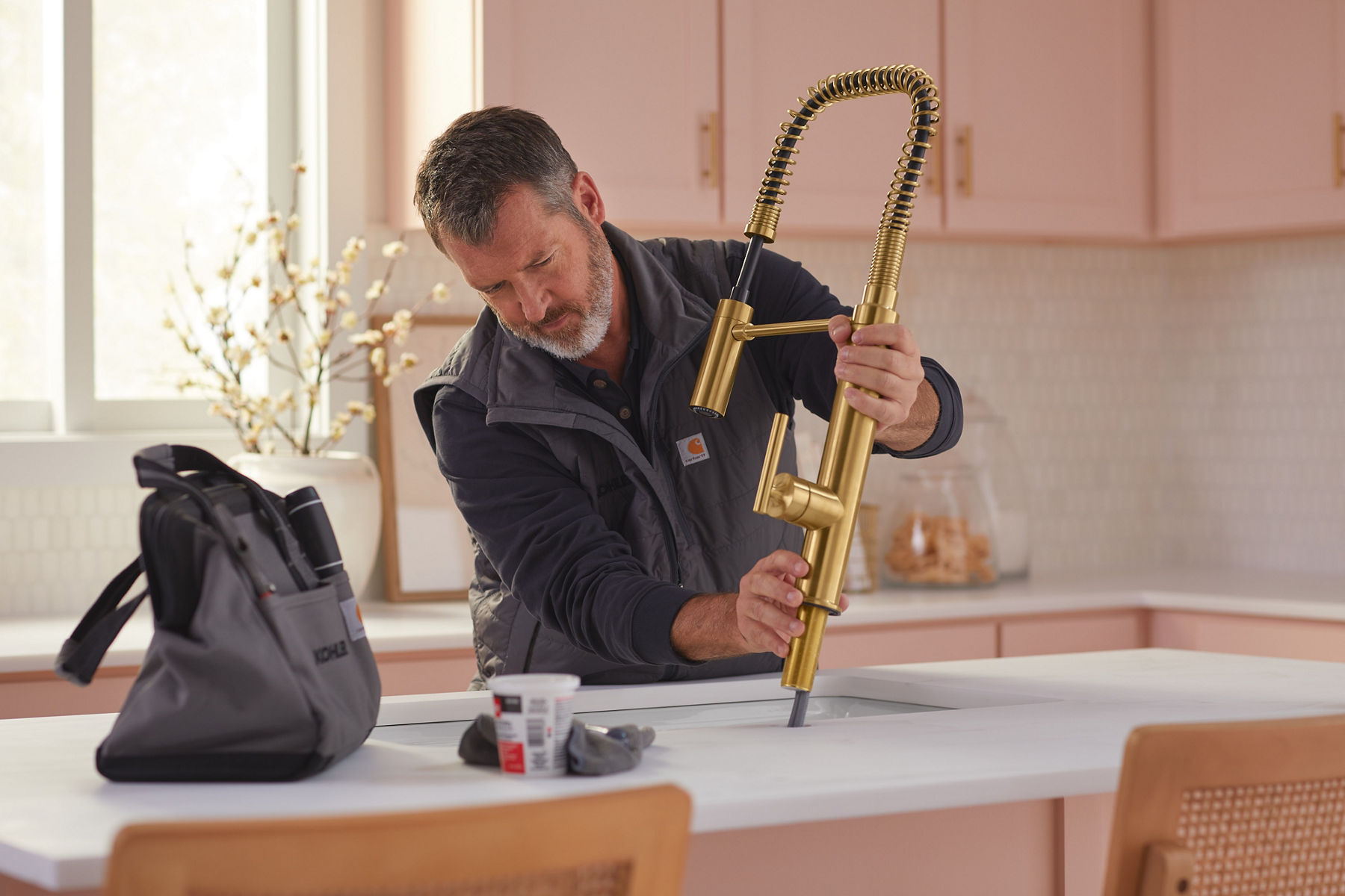 A man in work clothes installing a brushed brass faucet in a kitchen with pink cabinets and white counter tops.