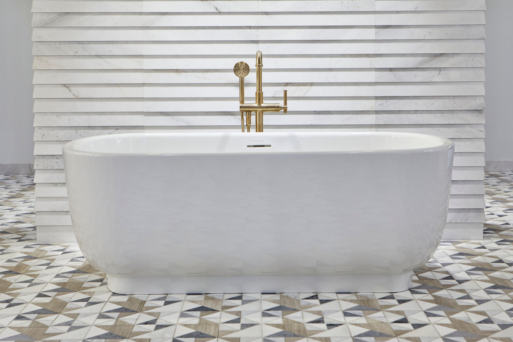 A large empty room with a patterned tile floor, a white freestanding bath with brushed brass bath filler in the center, an off-white stone wall behind it with the Ann Sacks, Kallista, and Robern logos on the wall.