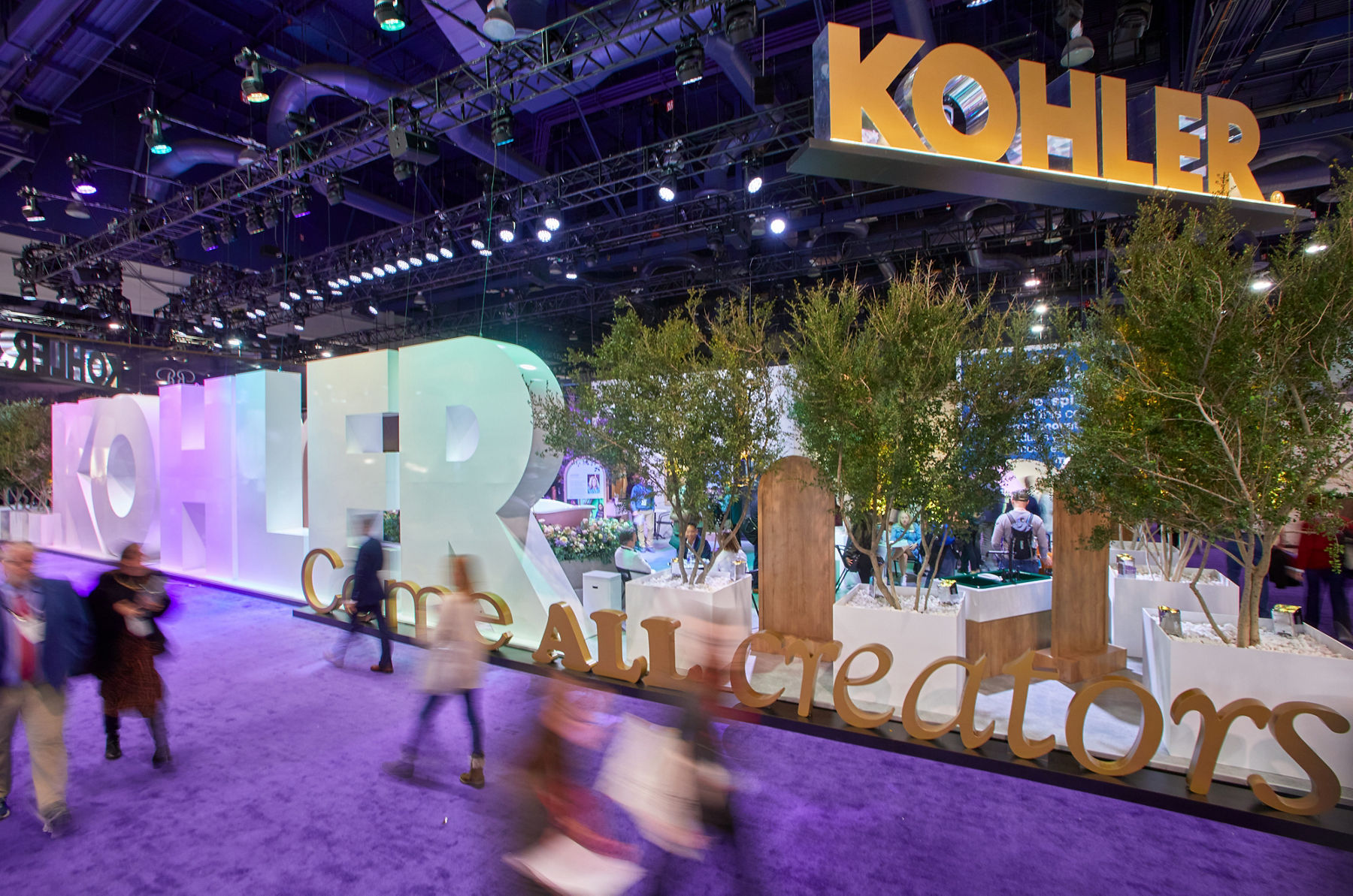 A Kohler tradeshow booth with the word KOHLER in large white three-dimensional lettering, roughly ten feet tall, and the words  Come All Creators in smaller three-dimensional lettering in three different type faces, roughly three feet tall, with several trees and displays behind them as people walk past in a slow shutter speed blur.