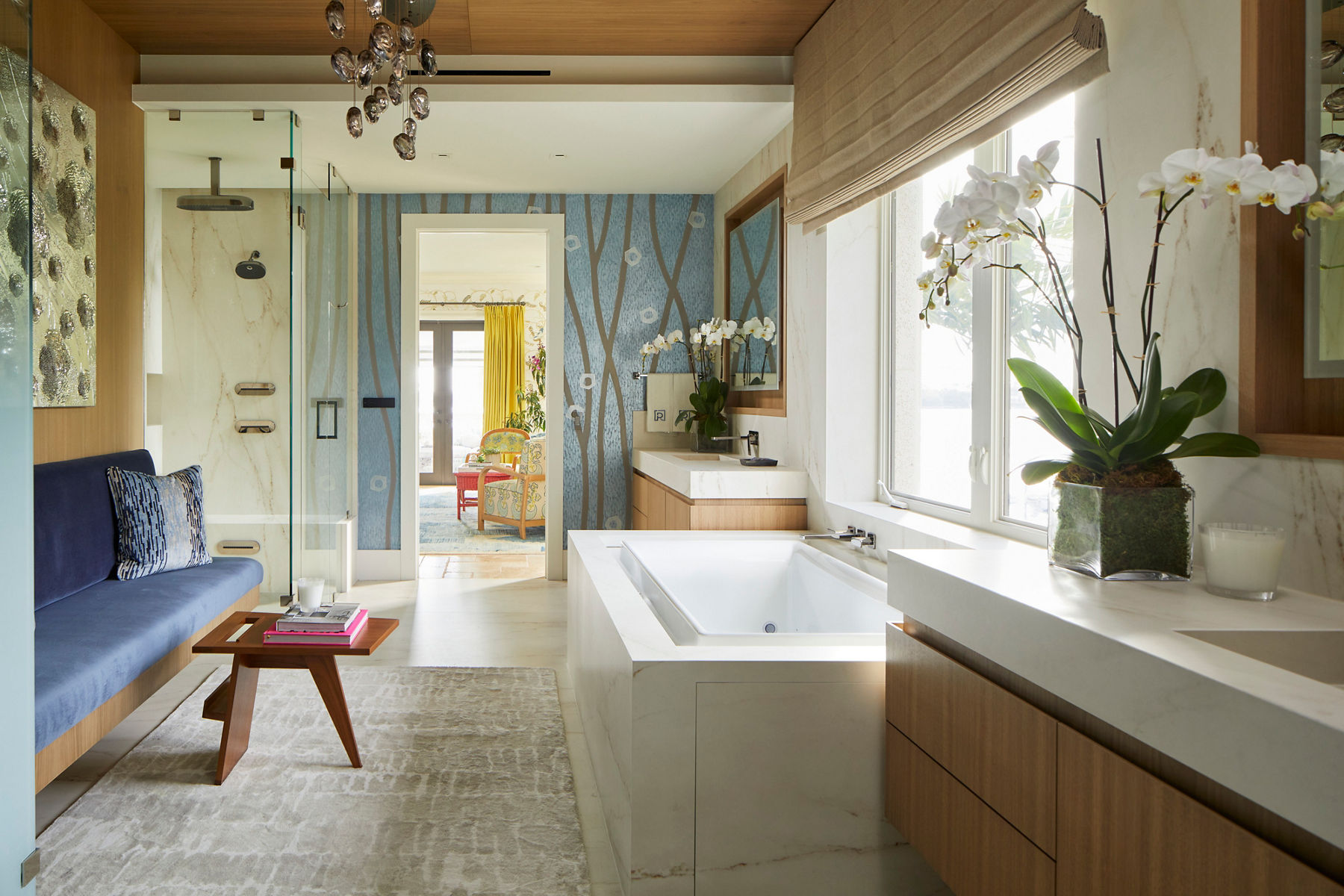 Wide image of a large bathroom space with a blue couch and coffee table across from a large white bath under a picture-frame window. There is a shower enclosed in glass with a rain shower head extending from the ceiling. The sink and vanity is in distance next to a blue accent wall with a pattern resembling tree branches. Through the doorway of the bathroom, you can see the primary suite and windows overlooking a body of water.