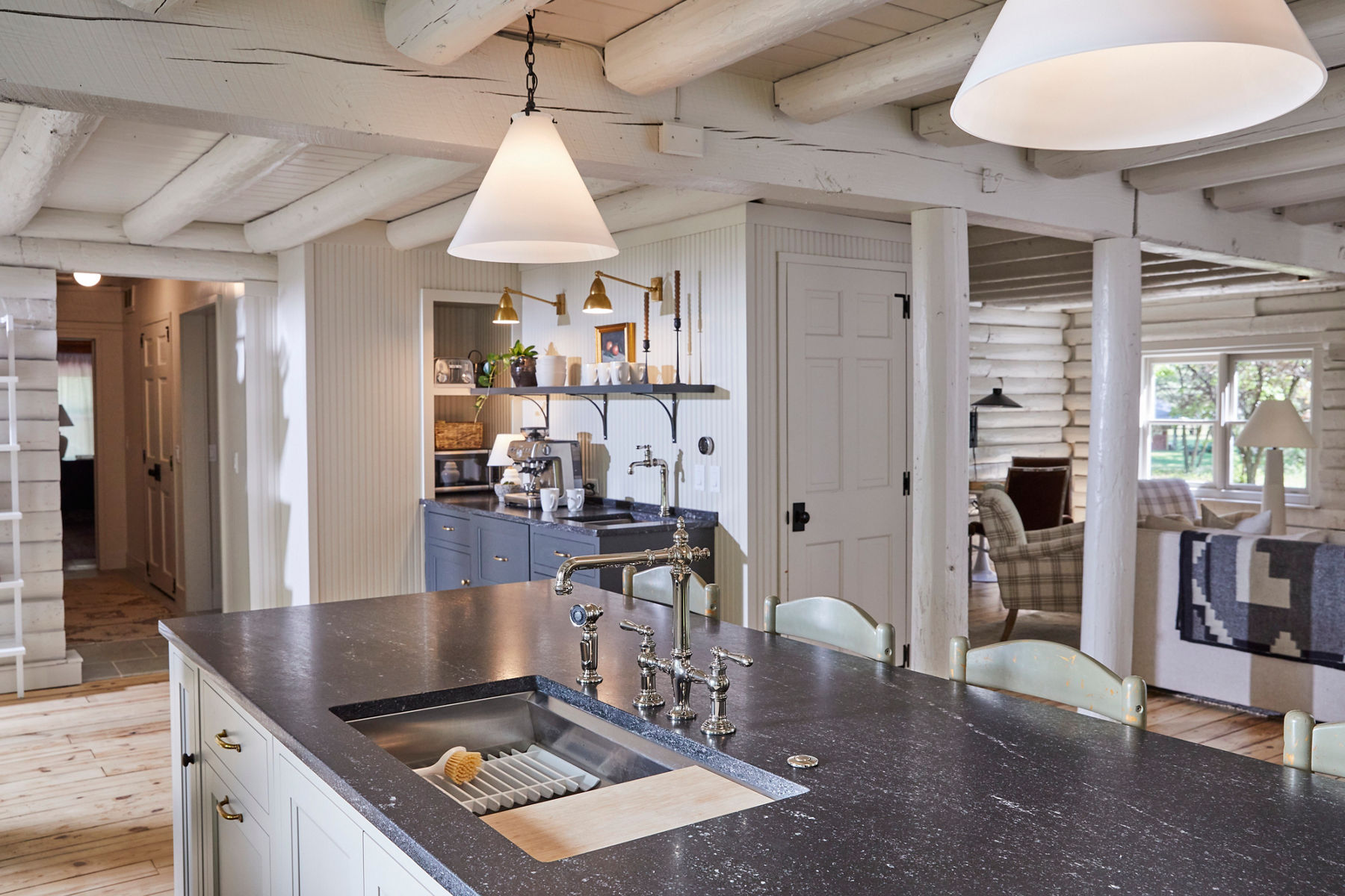 Wide image of the kitchen and coffee bar space in the fully renovated rental cabin designed by mother-daughter design duo Jean Stoffer and Grace Start. The kichen island has a dark stone counter top with undermount stainless steel Kohler workstation sink in the center with polished nickel Artifacts faucet. Coffee bar features a Kohler beverage sink and single-handle artifacts faucet in matching polished finish.
