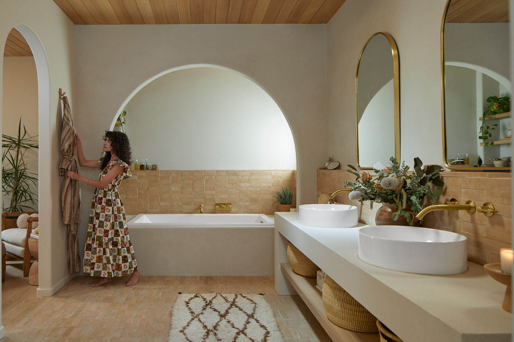 Wide image of a primary bathroom designed by Robert and Christina Martinez, the creative duo known as New Darlings. The bathroom features two round white vessel sinks and wall-mounted brass faucets. At the center of the image is the large soaking bath, placed next to a window to allow the Arizona desert sunlight to come in. Christina is in the image hanging a towel robe on a hook next to the bath. 