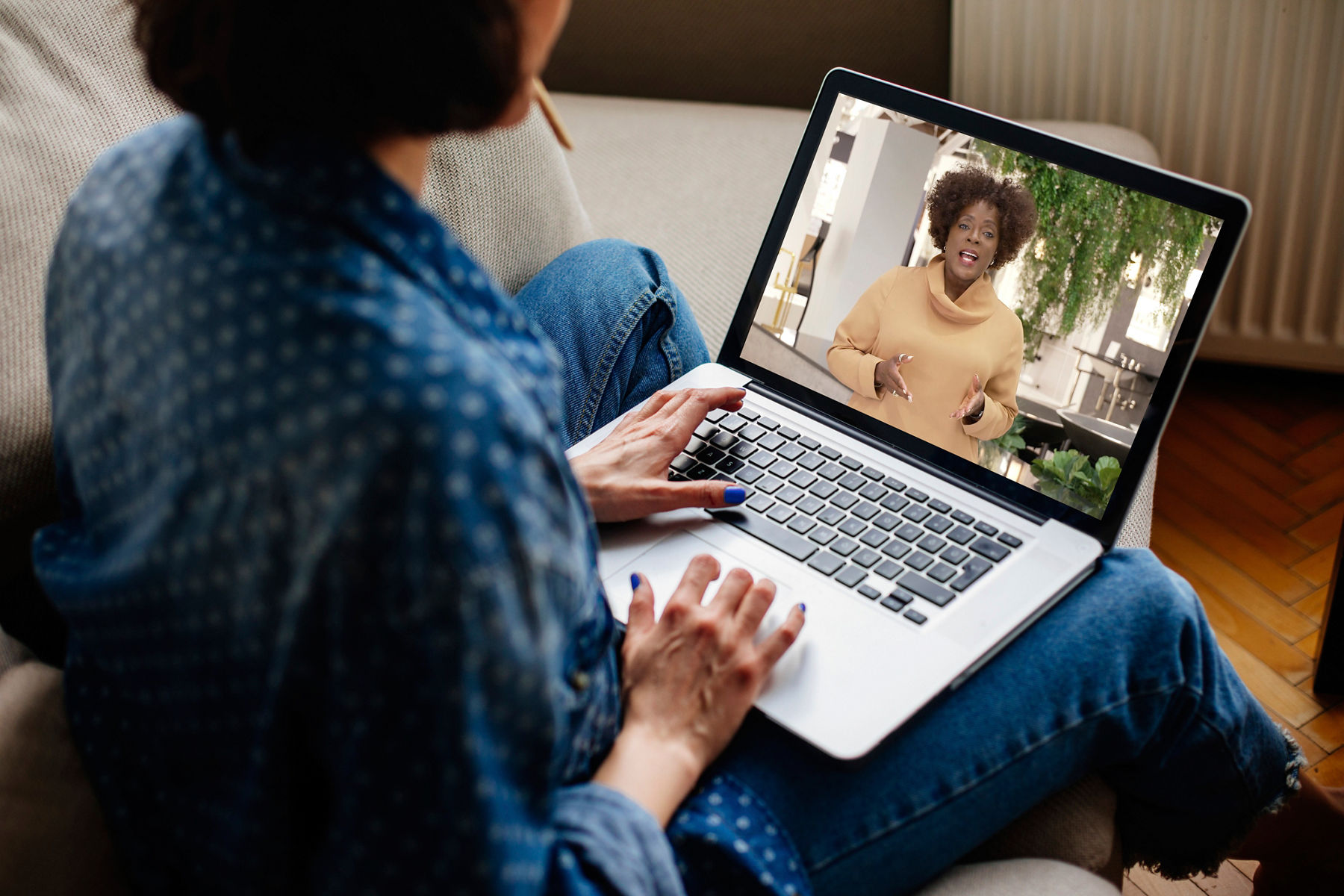 Image of a person casually dressed at home, watching a video on their laptop computer. Looking over their shoulder, the screen of the laptop shows a woman speaking in the video. 