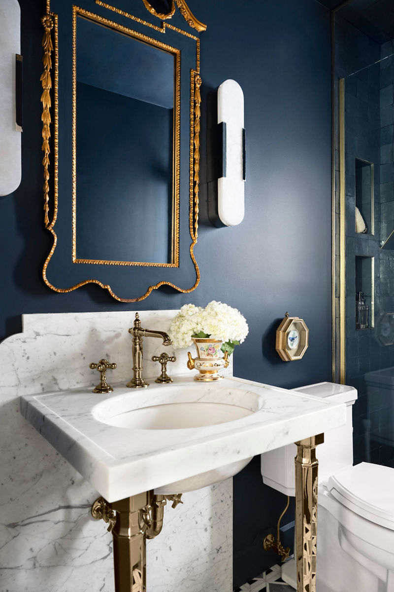 A landscape image of an elegant guest bathroom featuring deep blue walls, a white marble countertop with polished metal fixtures, a decorative vase with white hydrangeas, and a gold-framed octagonal mirror.