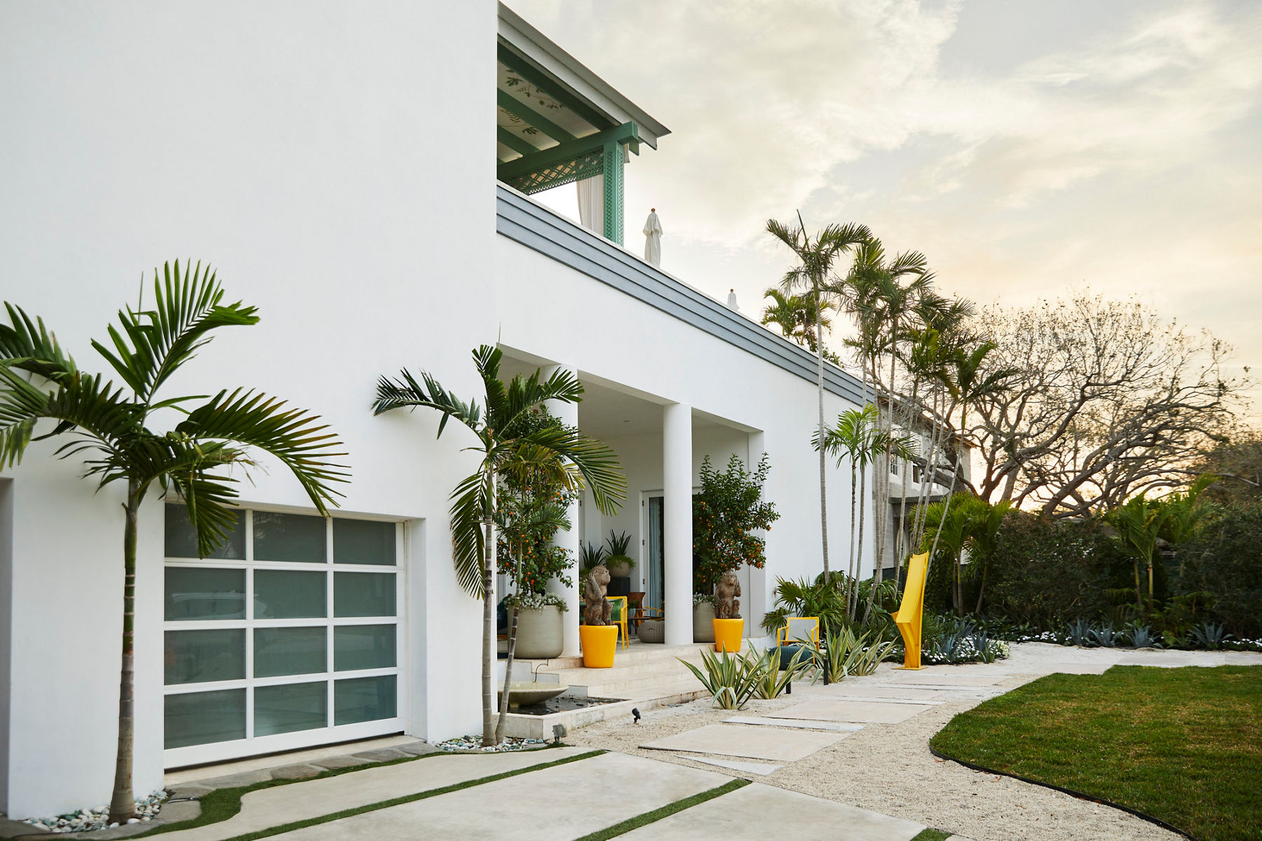 Exterior of the Kips Bay Palm Beach show house with a white building, bright yellow sculptures and planters, and abundant tropical plants.