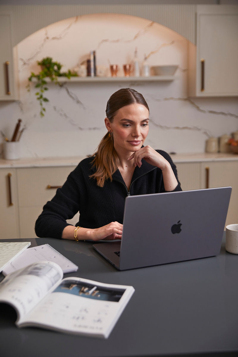 A woman with light brown hair tied back in a ponytail is sitting at a modern kitchen counter, focused on her work on a gray Apple MacBook. She is dressed in a black sweater and wearing gold hoop earrings and a gold bracelet. The countertop has a notebook and an open catalog or magazine, while a coffee mug sits nearby. The kitchen in the background features white cabinets with gold handles, a marble backsplash with subtle veining, and open shelving with neatly arranged items, including a small potted plant.