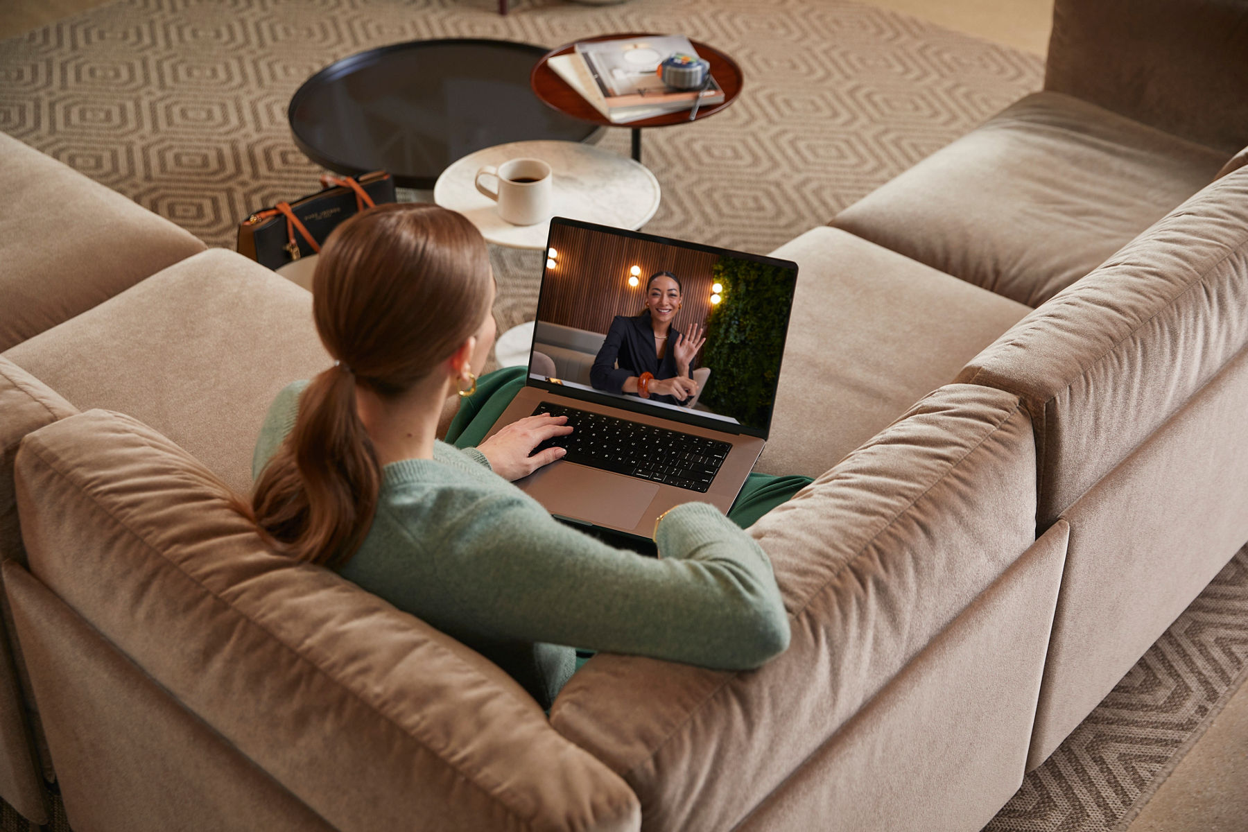 A woman with light brown hair tied back in a ponytail is sitting on a beige sofa, engaged in a video call on her laptop. She is wearing a light green sweater and gold hoop earrings. The laptop screen shows a woman smiling and waving from a warmly lit, modern space with wooden paneling and green plants in the background. In the foreground, there is a small round coffee table with a cup of coffee, a stack of books, and a pair of eyeglasses. The setting appears cozy and comfortable, with a patterned carpet and additional seating in the background.