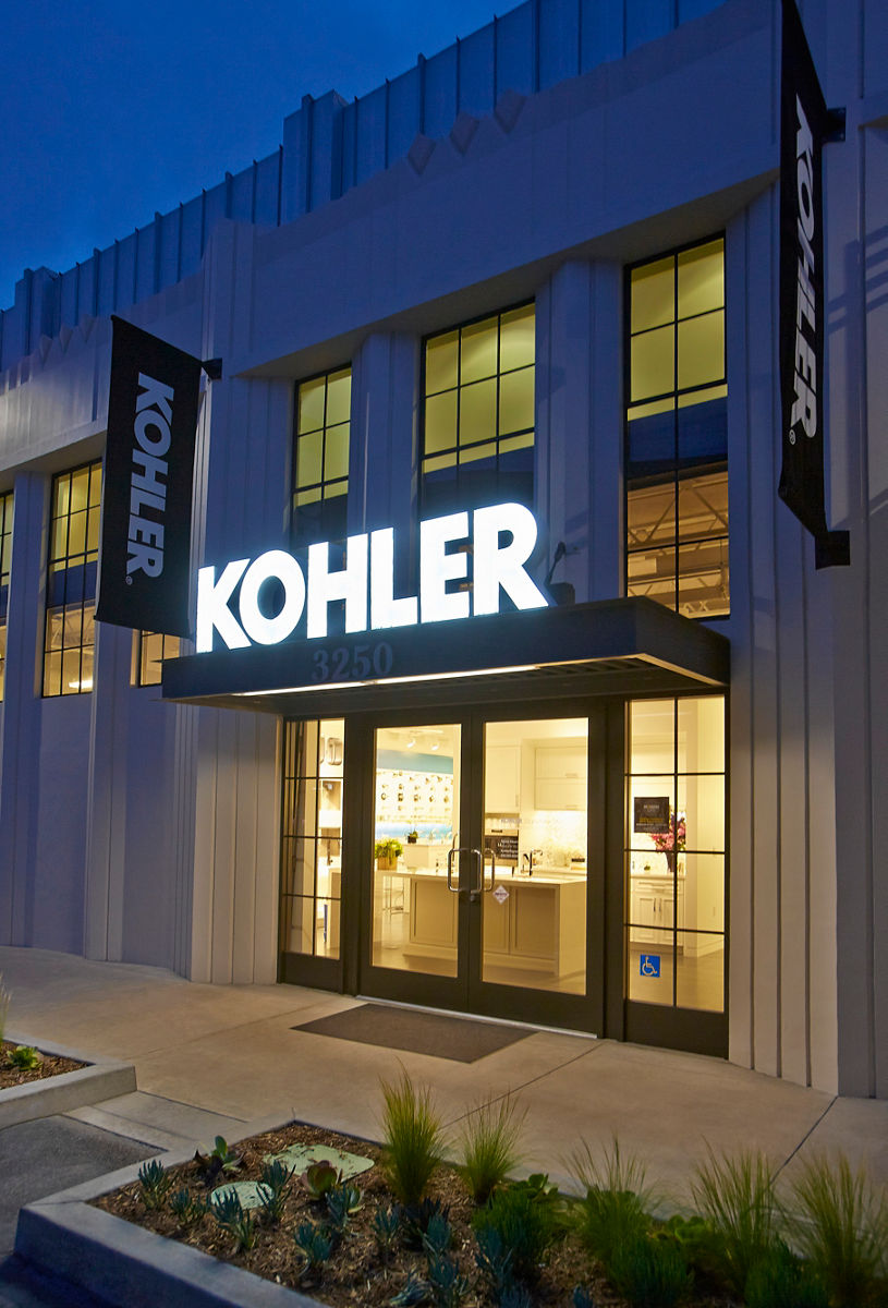 Exterior view of a Kohler store at dusk, with a brightly illuminated 'KOHLER' sign above the entrance. The building has a modern design with tall windows and vertical architectural details. Two black banners with the white Kohler logo hang on either side of the entrance. The glass doors reveal a well-lit interior showcasing bathroom and kitchen fixtures.