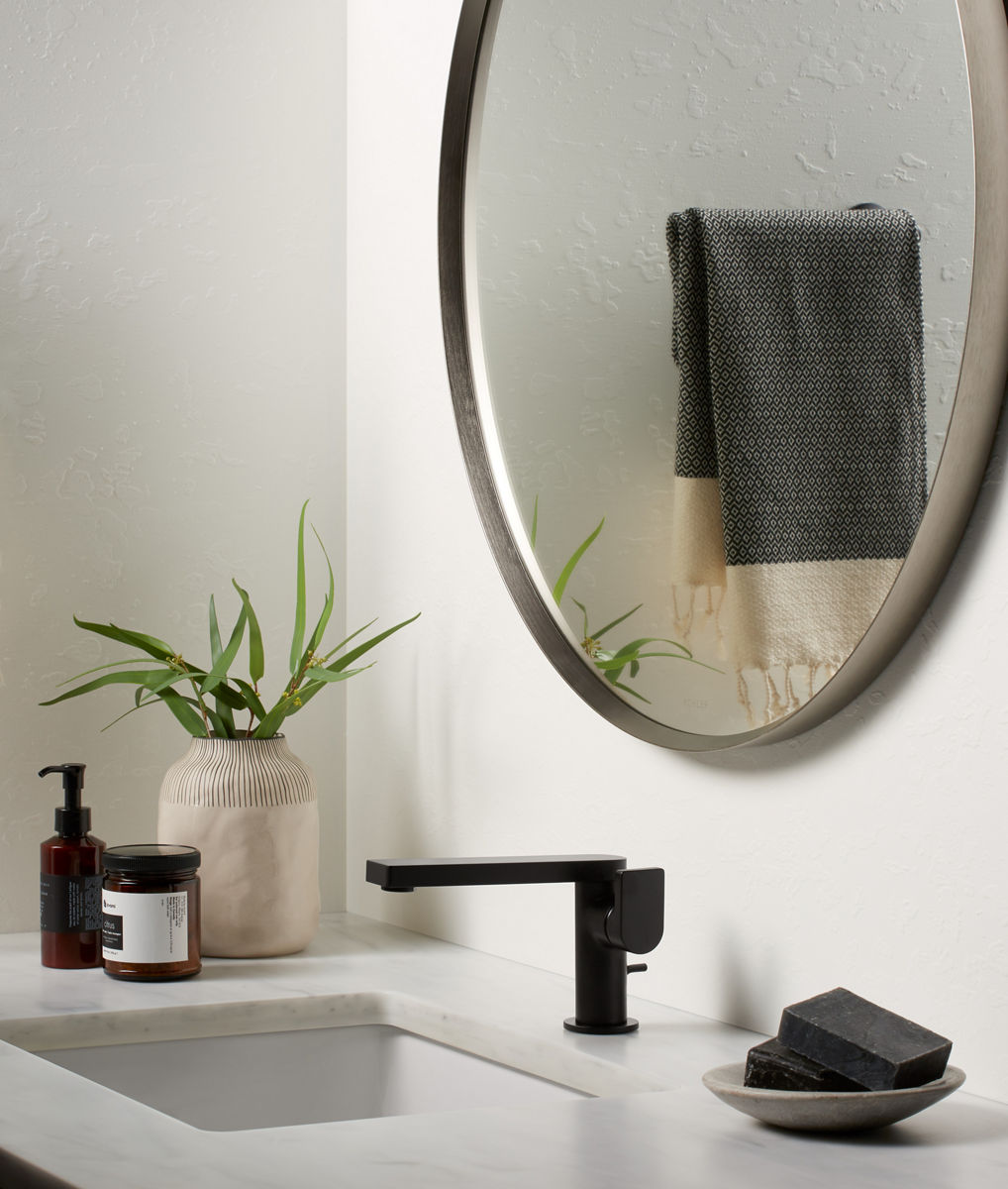 A round, metal-framed mirror against a white wall over a white vanity counter top with a black faucet and white undermount sink.