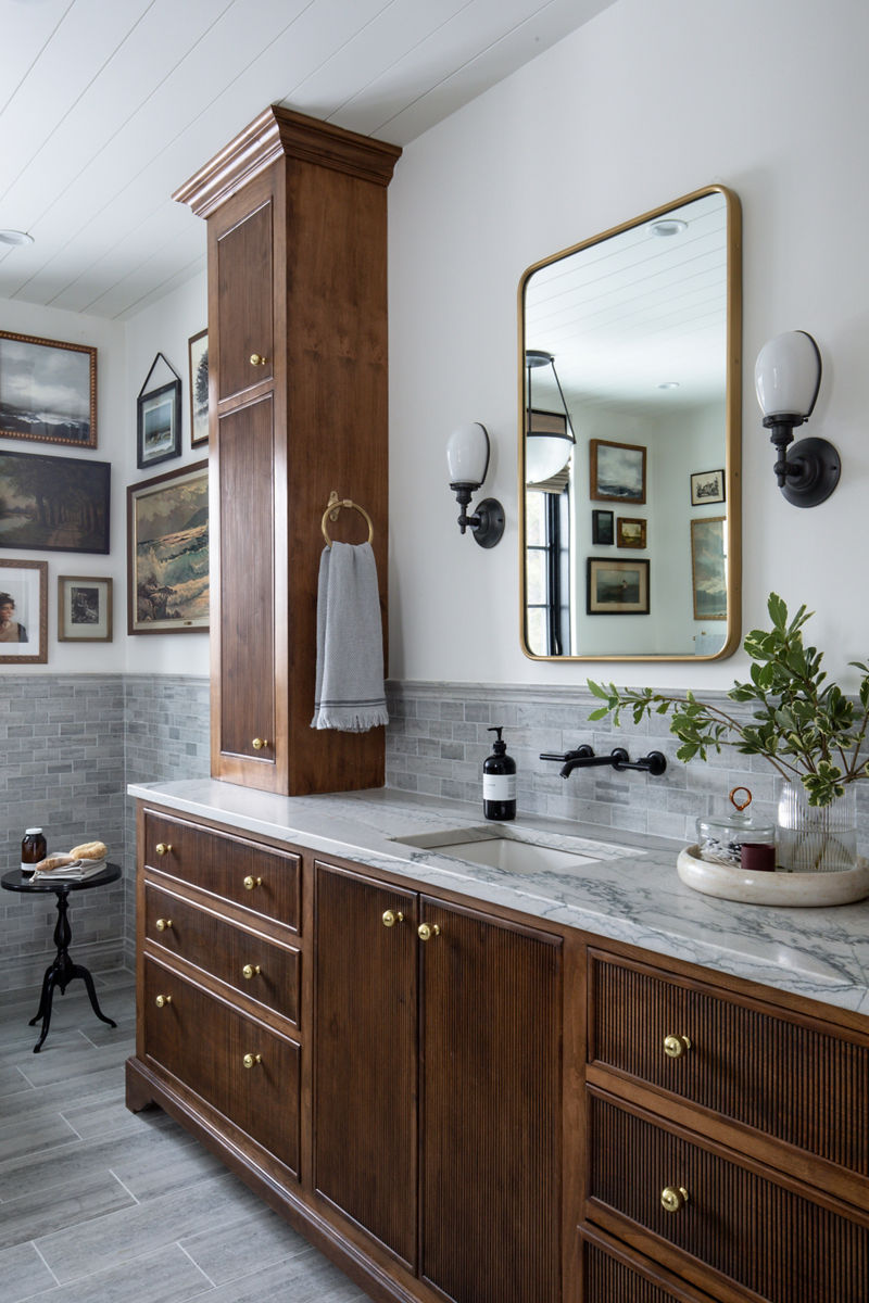 Une salle de bains au design classique avec un dosseret carrelé, un robinet mural déployé KOHLER avec des poignées à levier en noir mat, un dessus de meuble-lavabo en marbre, ainsi qu’un meuble-lavabo et une armoire à linge avec des garnitures traditionnelles et des détails cannelés.