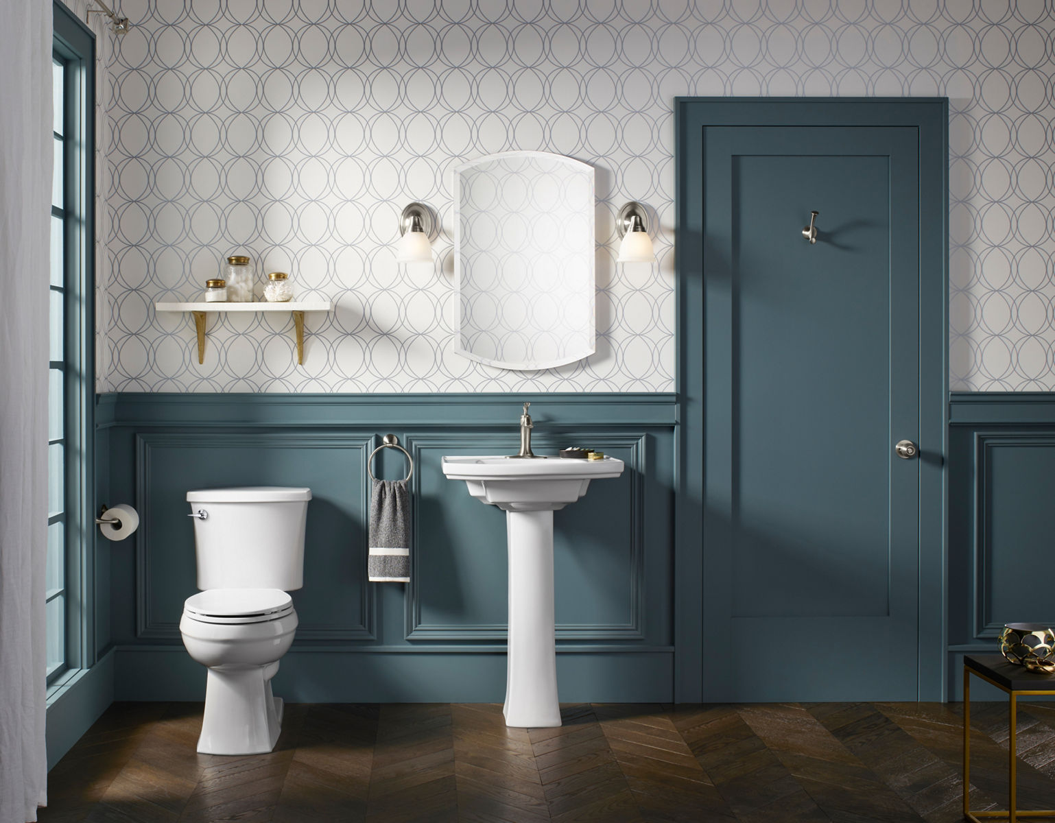 Une armoire à pharmacie sans cadre KOHLER est suspendue dans une salle de bain nouvellement conçue à côté de deux appliques à ampoule unique, une étagère murale, un lavabo sur colonne et une toilette KOHLER.