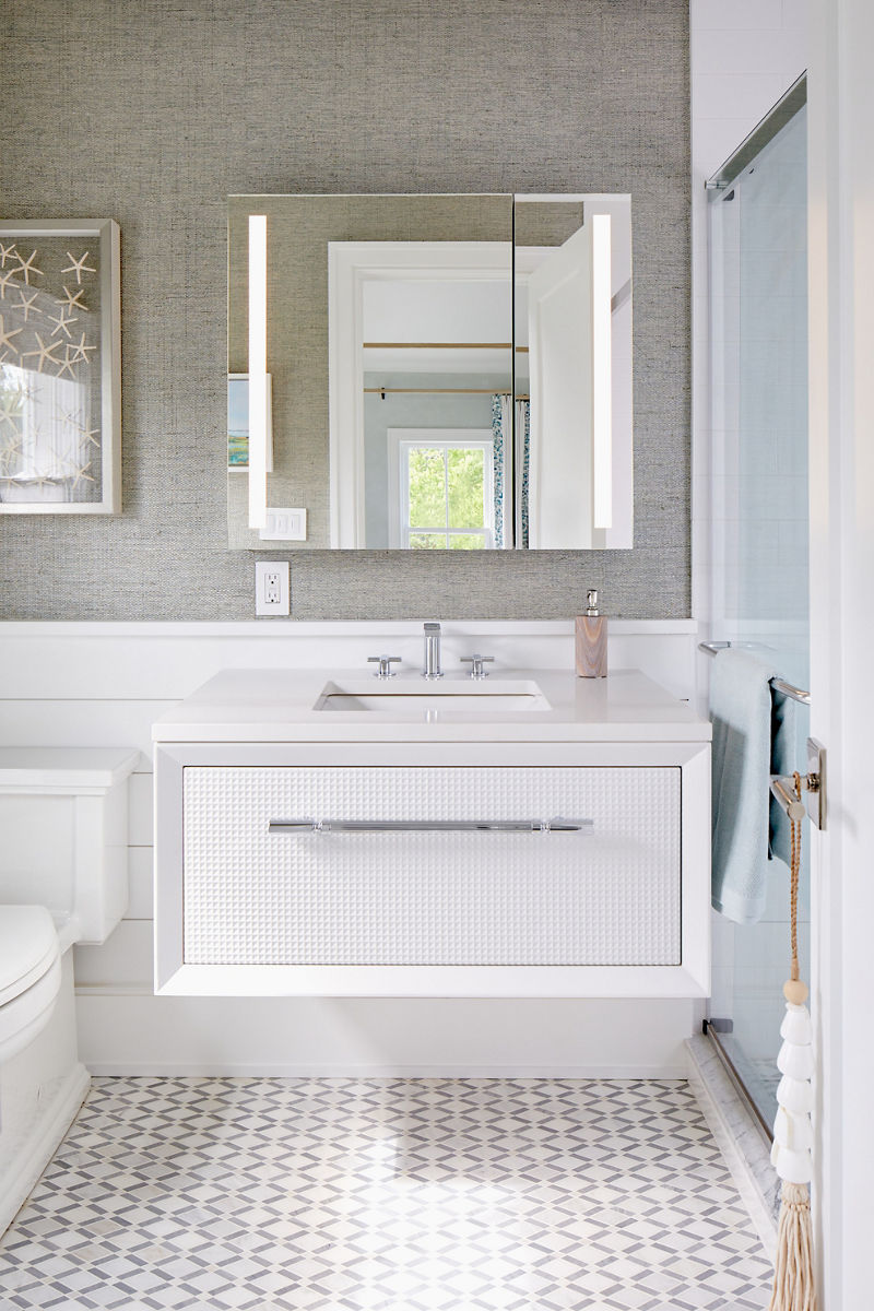 A lighted mirror against a textured gray wall above a whtie vanity with chrome faucet and hardware.