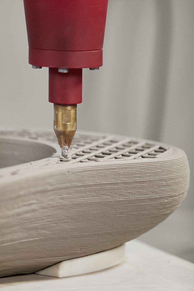 Gros plan sur un évier Rock.01 imprimé en 3D. La buse d’impression applique le matériau de porcelaine vitrifiée sur un évier partiellement rempli, montrant la structure de renfort interne et les couches extérieures du processus d’impression.