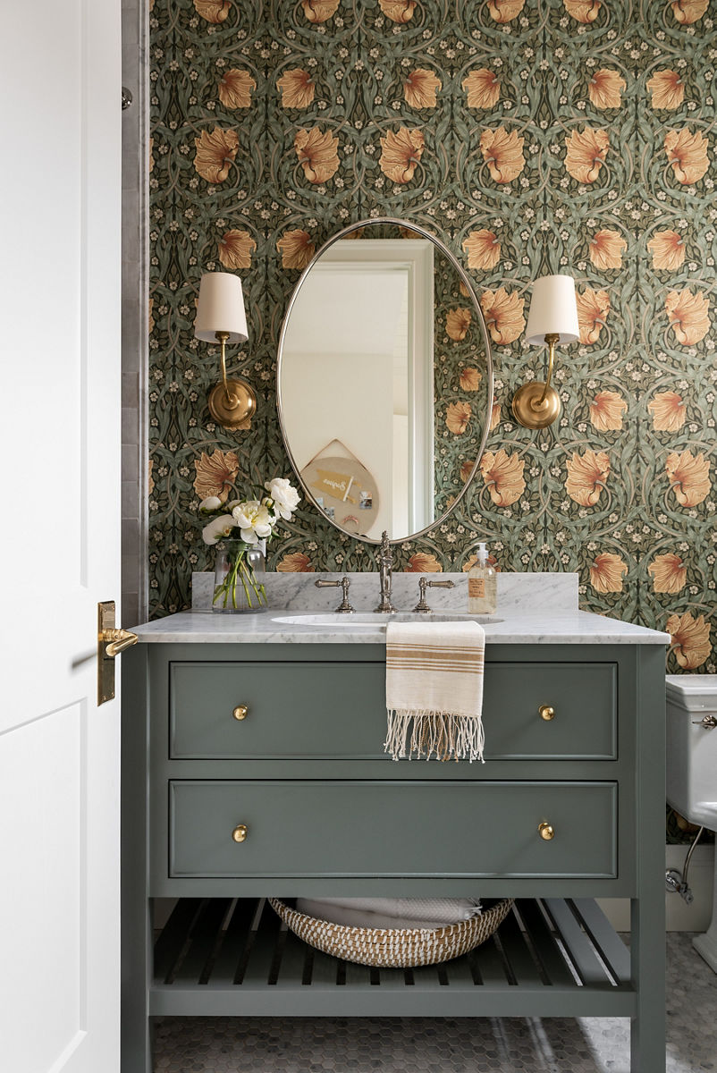 A Grandmillennial style bathroom with white topped sage green vanity, KOHLER widespread lever handle faucet, sconces with white shades, oval mirror, and floral patterned wallpaper.