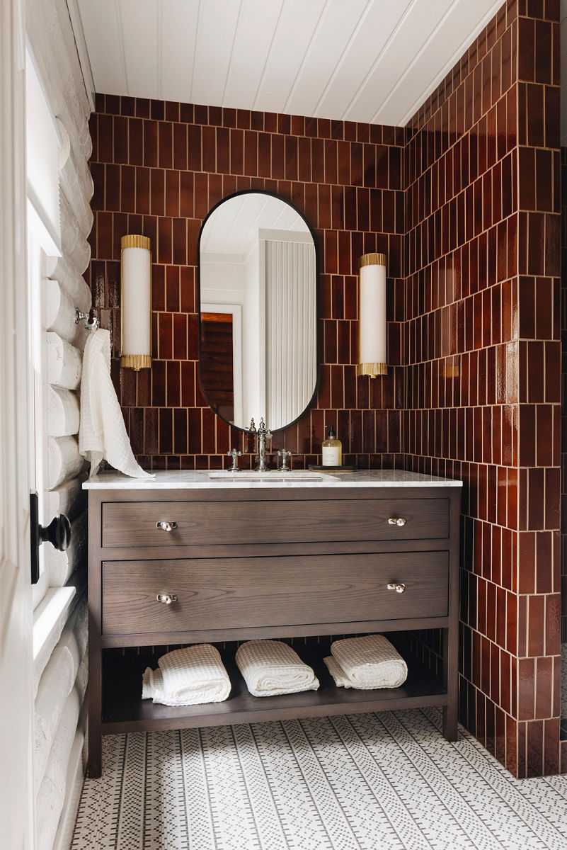 Image of the vanity space in the cabin bathroom where a rustic wood freestanding vanity with open towel shell on the bottom sits in front of a bold rootbeer-colored glossy tile wall from floor to ceiling. A Kohler Artifacts bathroom faucet in polished nickel finish is atop the white counter top, with vertical sconses on either side of the oval matte black rimmed mirror.