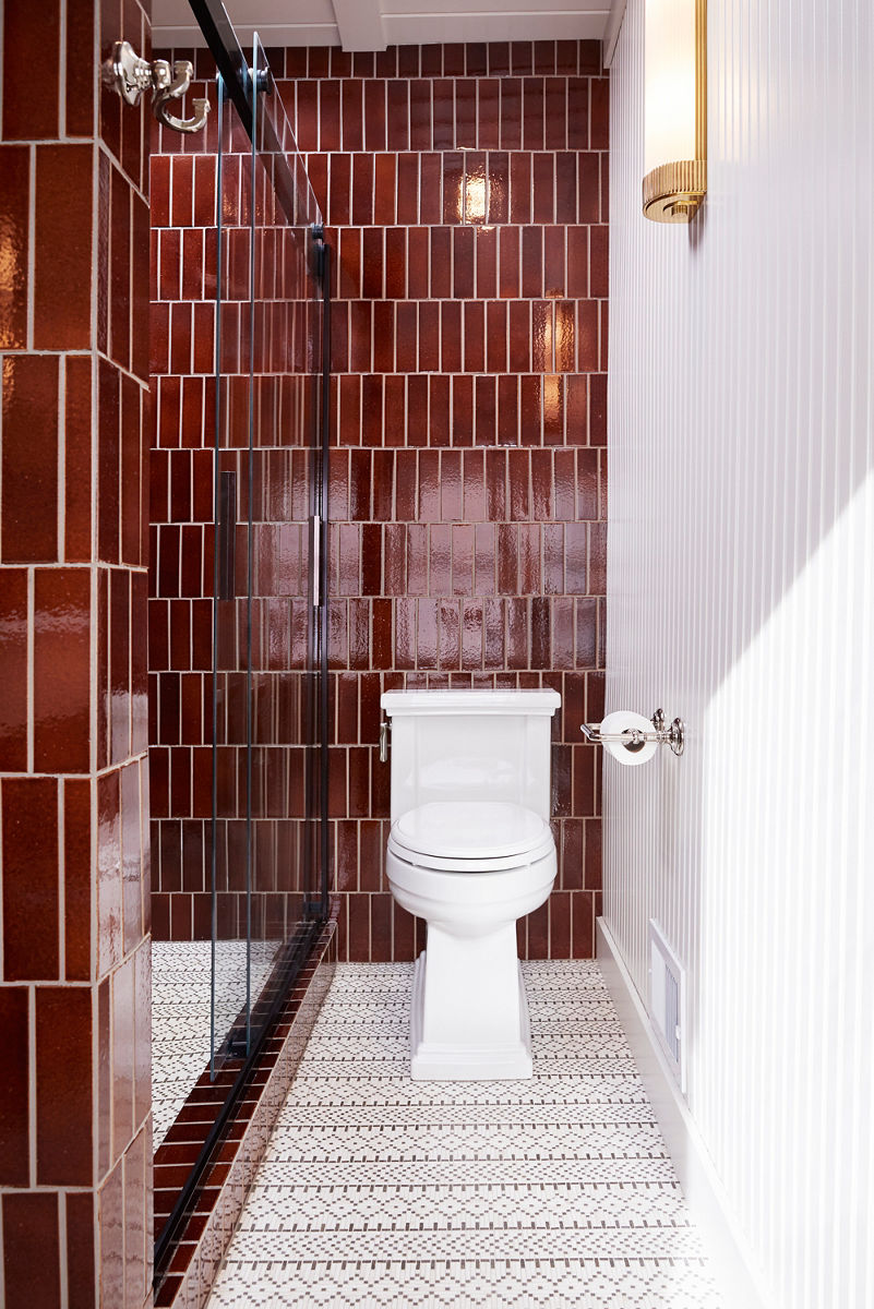 Image of the white Tresham toilet centered in front of the vertically tiled, rootbeer-colored wall and intricate geometric patterned white and black floor tile. The toilet paper holder on the wall is of polished nickel finish.