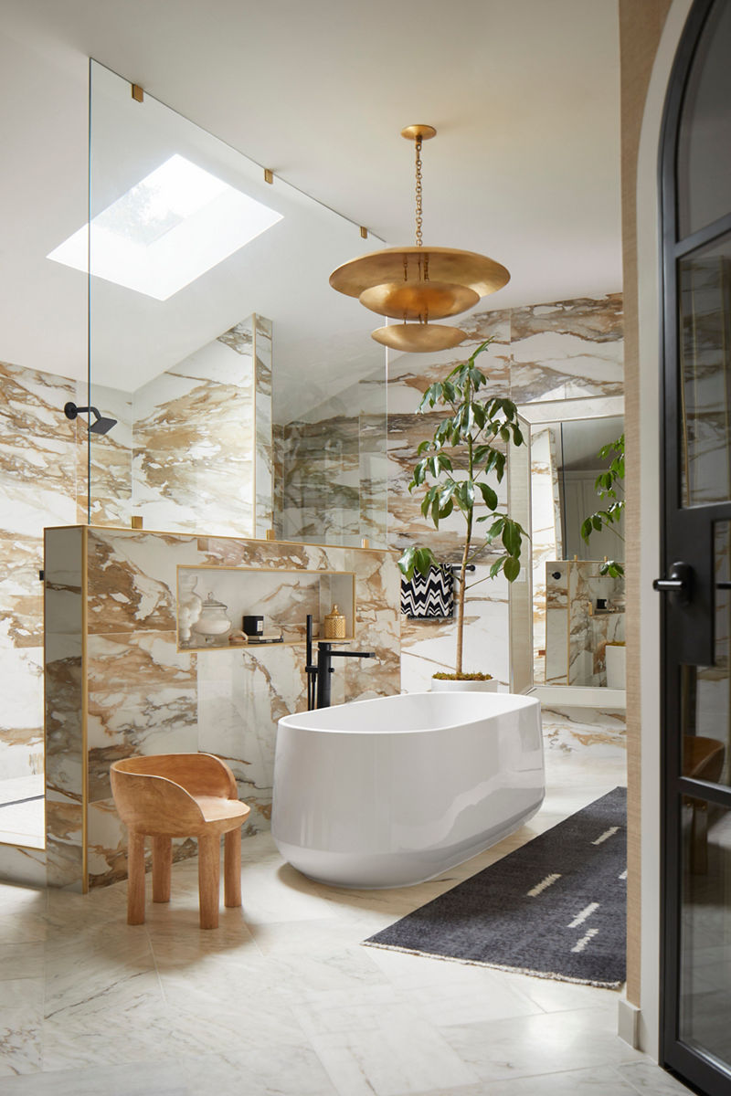 Image of Nikki Chu's primary bath suite featuring a white, oval Kohler freestanding bath as the centerpiece of the room. Above is a skylight in the vaulted ceiling and a gold chandelier. The tile walls have a gold and white marble tone, enclosing the shower area.