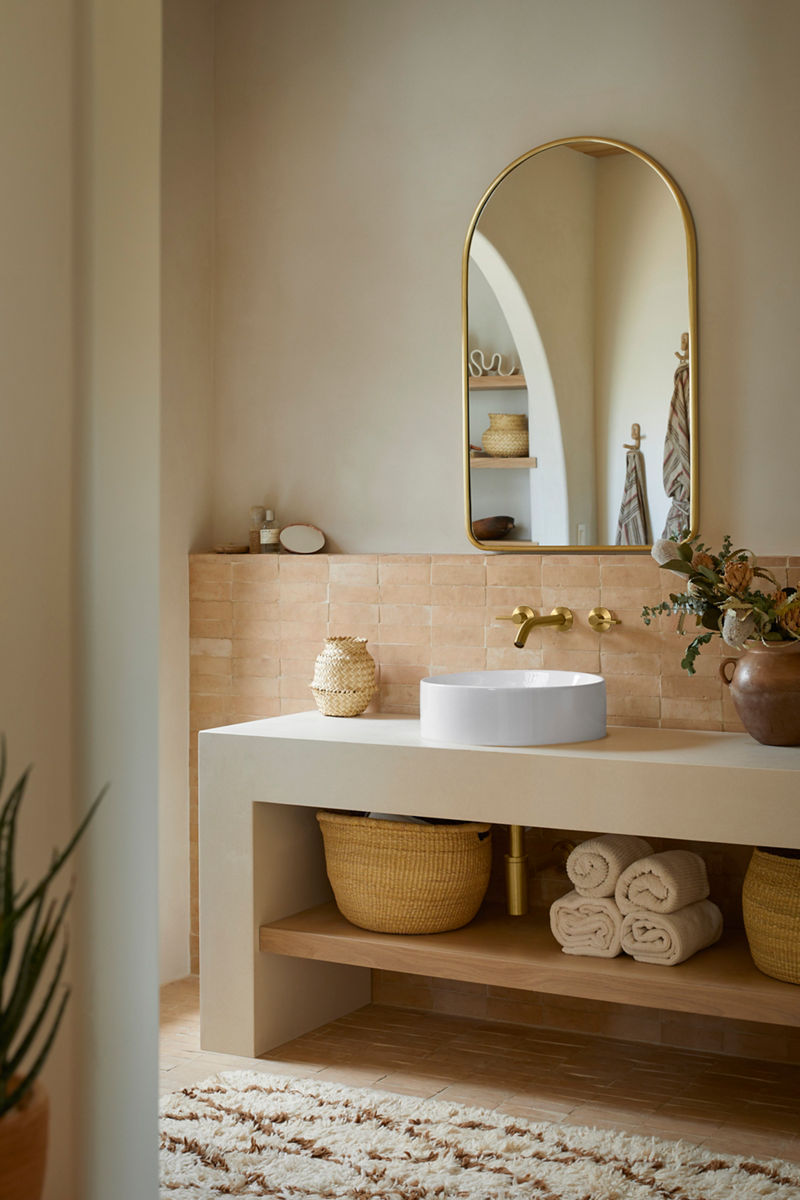 Image of the sink space in the primary bathroom featuring a white, round vessel sink and wall-mounted brass Kohler faucet, and above is an arched mirror with brass finish trim. 