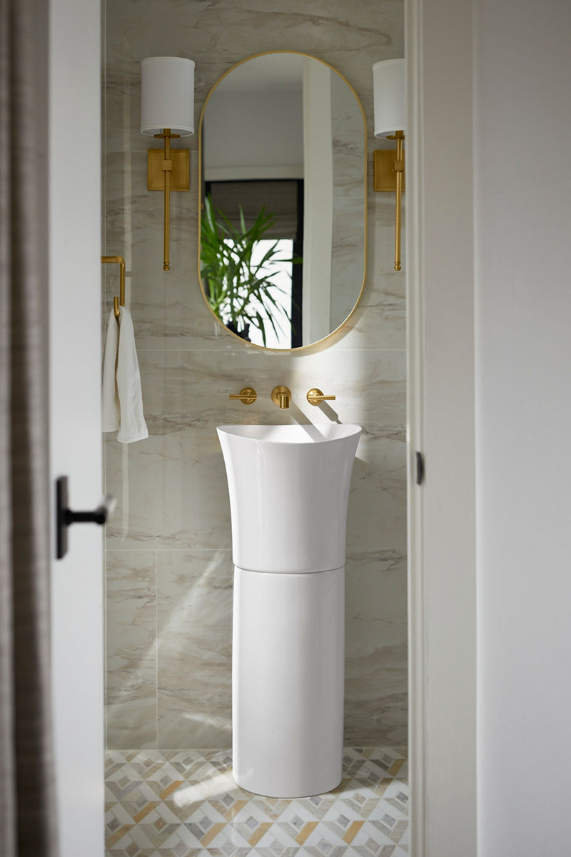 Image of Nikki's guest bathroom, centered on the wall is a vanity set up featuring the tall, white, oval pedestal sink from Kohler and a Kohler wall-mounted brass faucet. Above the sink and faucet are two matching brass sconces with white shades and an oval mirror with matching brass trim.