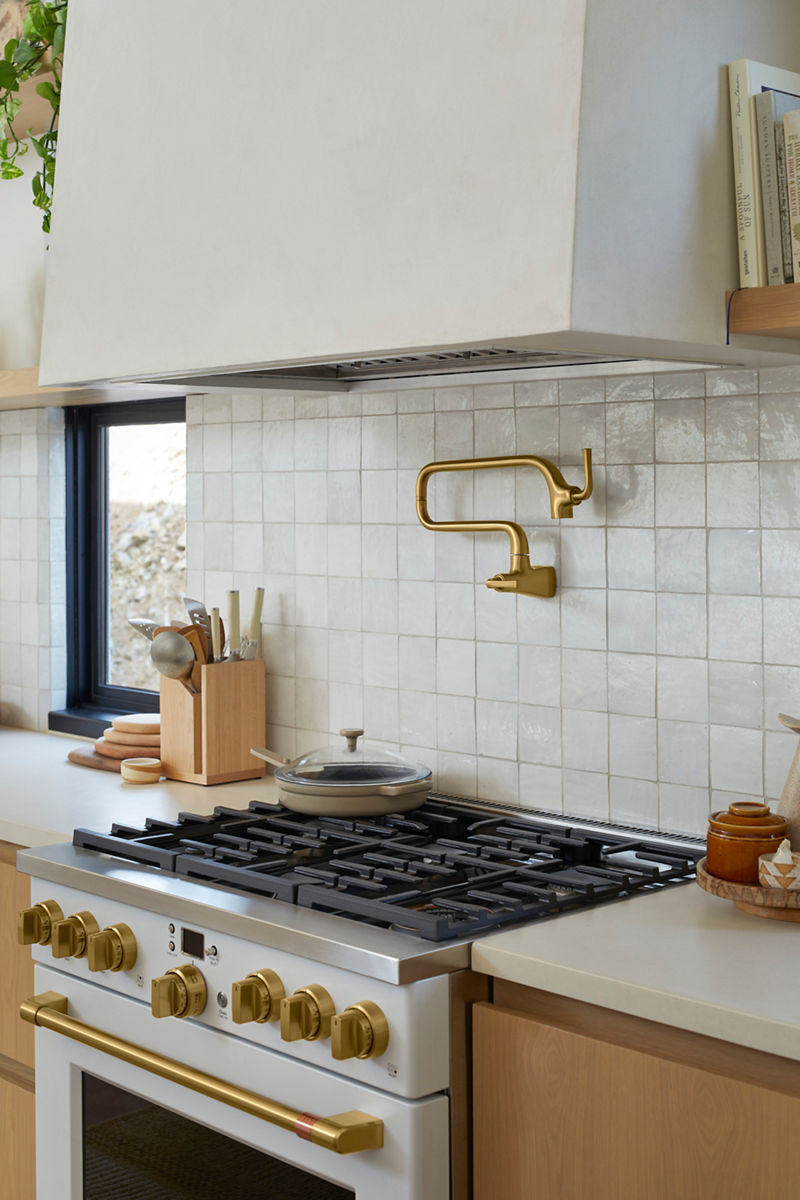 Image of the kitchen range area where a monochromatic soft white tile is featured as a backsplash under the range hood, and a Kohler brass pot filler faucet is centered on the wall.