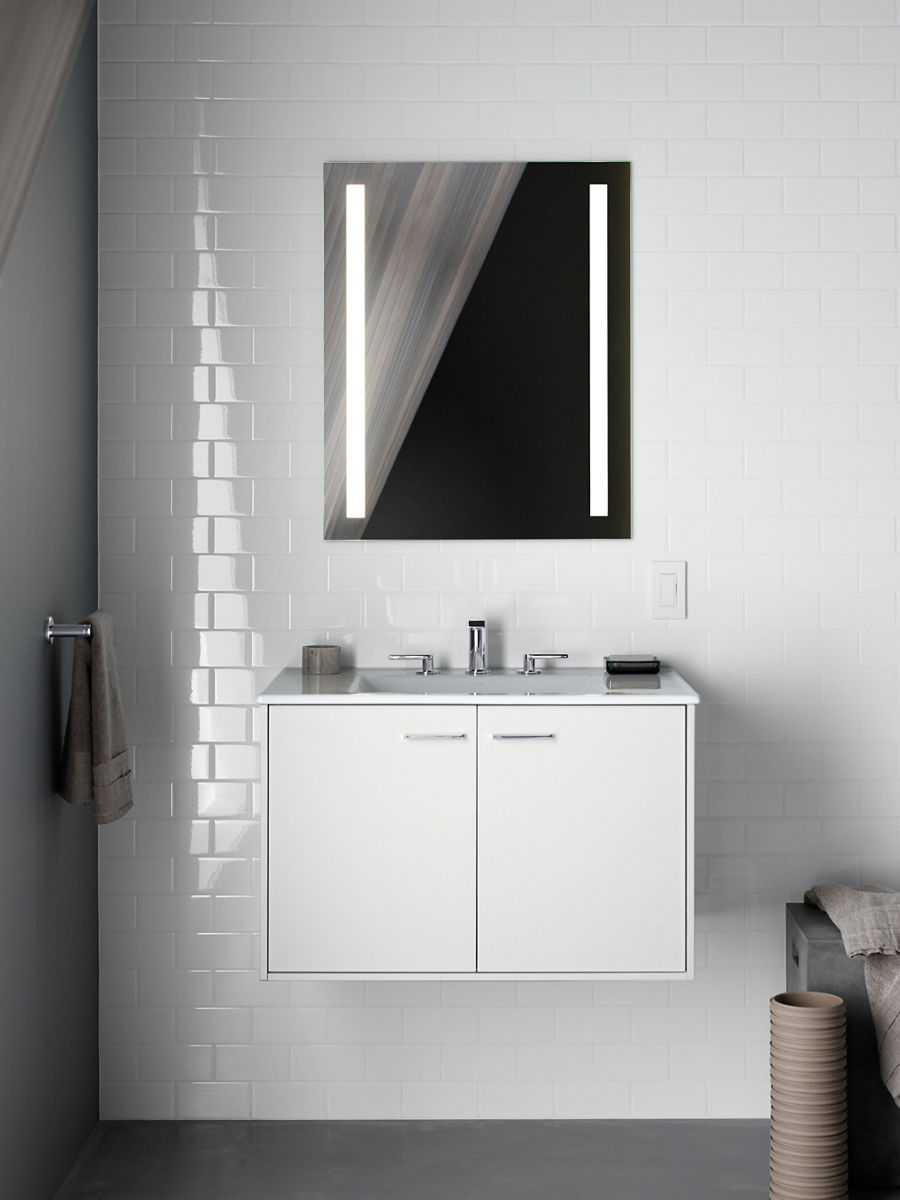 A lighted mirror hanging against a white tiled wall above a white vanity with a chrome faucet.