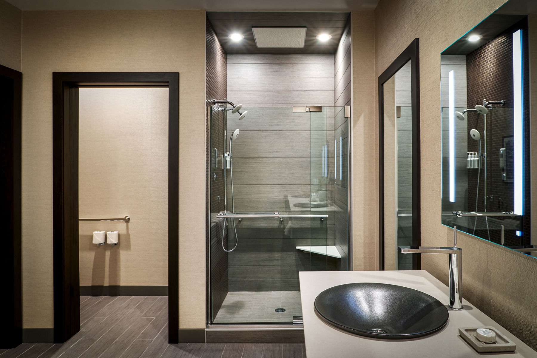 Interior view of hotel bathroom featuring illuminated shower stall with glass door fitted with Kohler shower trim.
