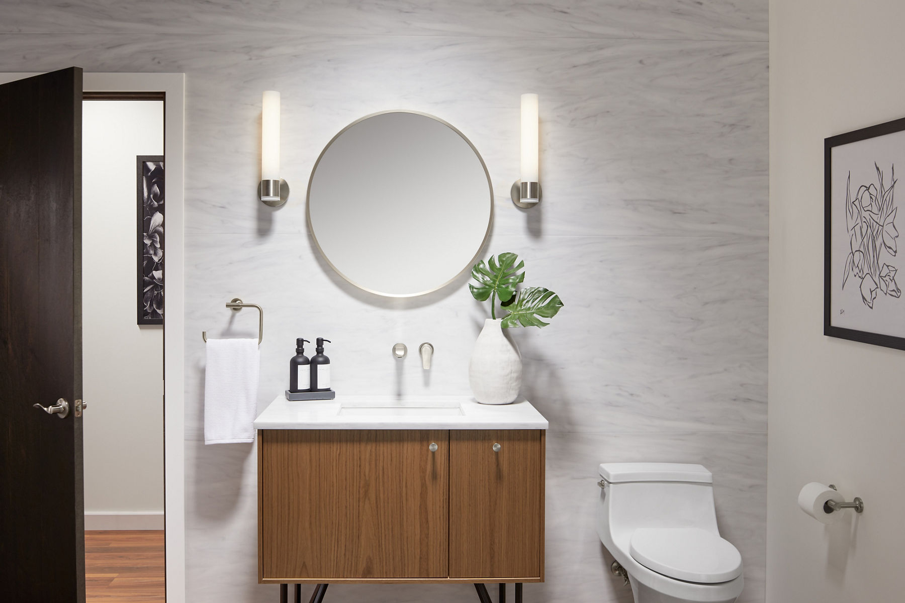 A brushed nickel-framed circular mirror with two sconce lights on either side on a white wall with a brushed nickel wall-mount faucet above a wood vanity with white counter and sink.