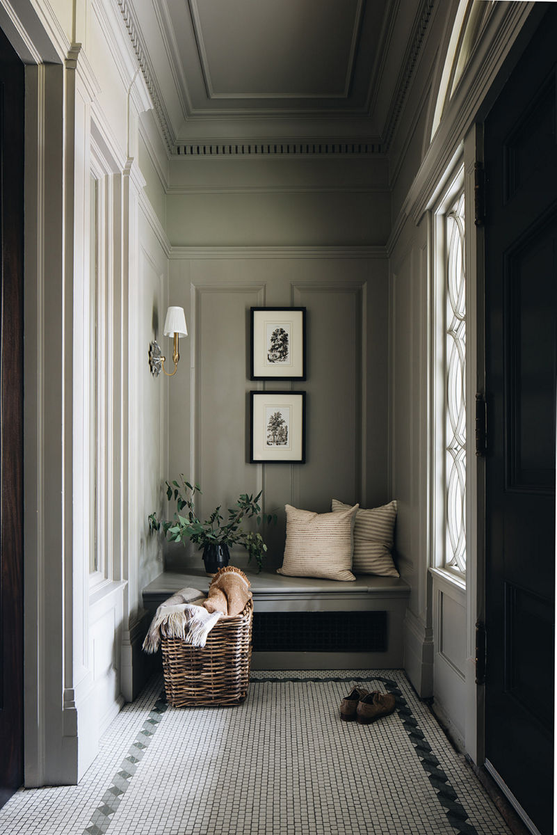 Light streams through a window in sage green paneled nook, with bench, penny tile mosaic floor, framed art, and a basket of blankets.