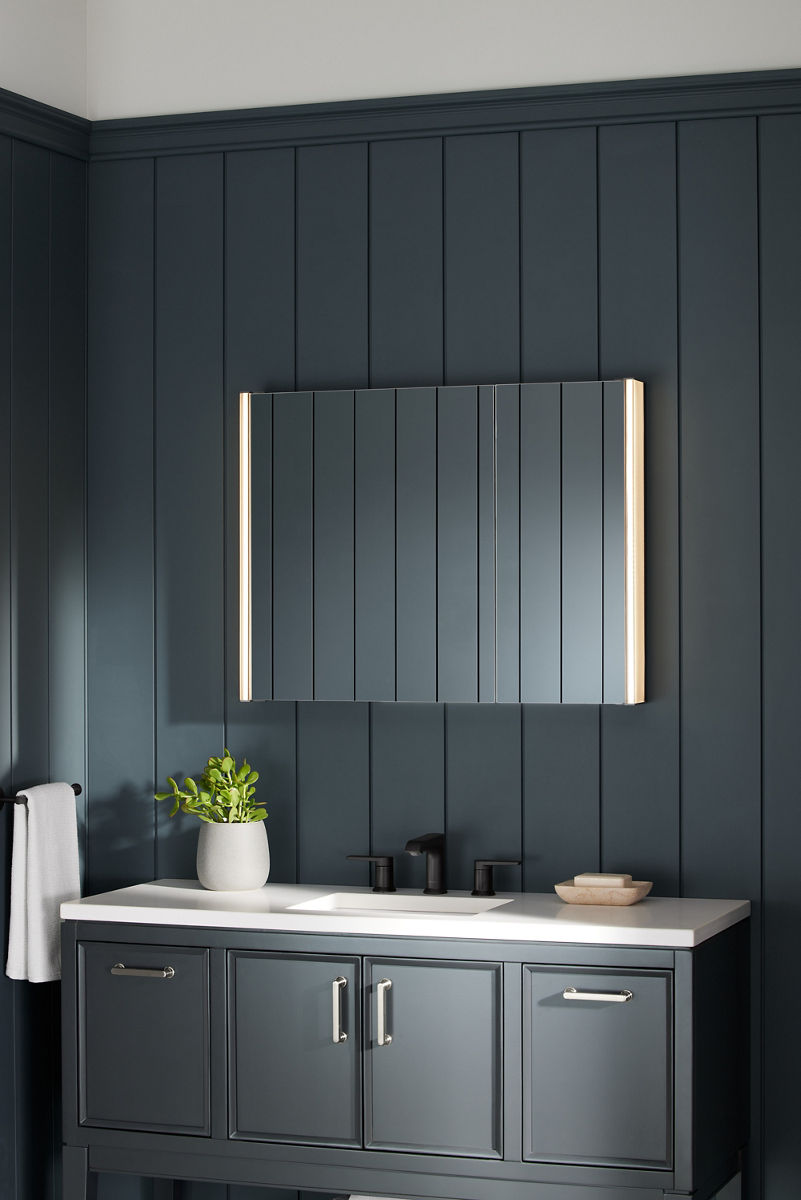 A KOHLER Verdera lighted medicine cabinet shines in a moody bathroom with black paneled walls, dark gray vanity, and white vanity top.