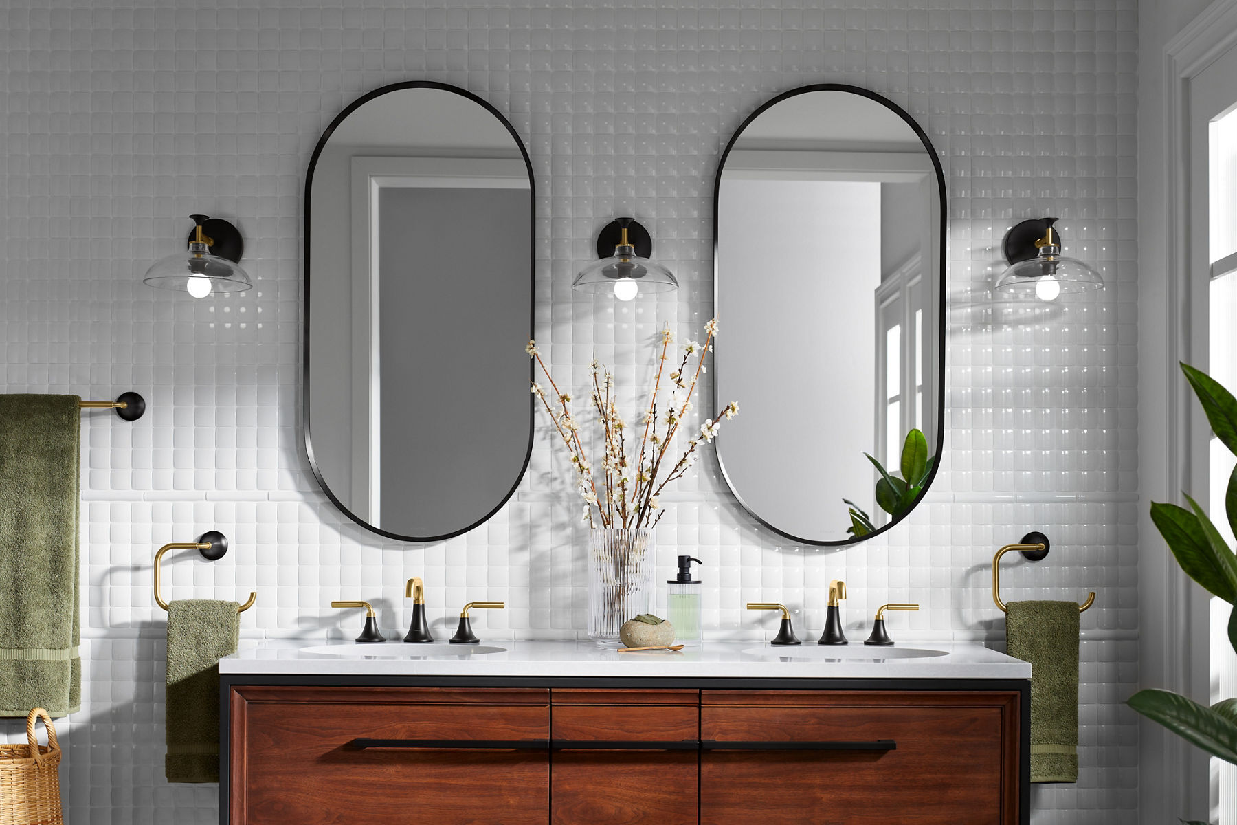 A sunlit bathroom with a large oval mirror, black and gold faucets, white tiled backsplash, and two wall-mounted glass lights, featuring a vase with flowers and a green towel.