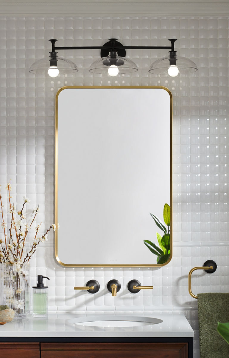 A gold-framed rectangular mirror with rounded corners against a white tile wall with a black, three-light sconce above it and a wall mount two tone faucet below.