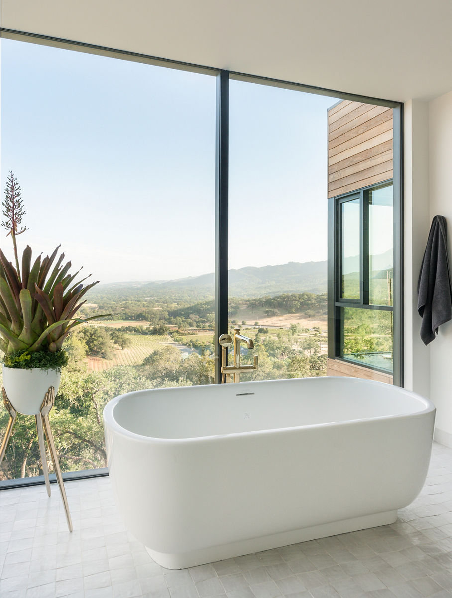 A Laura Kirar white freestanding bath in a bathroom with a floor-to-ceiling window overlooking a nature scene with small mountains in the distance.