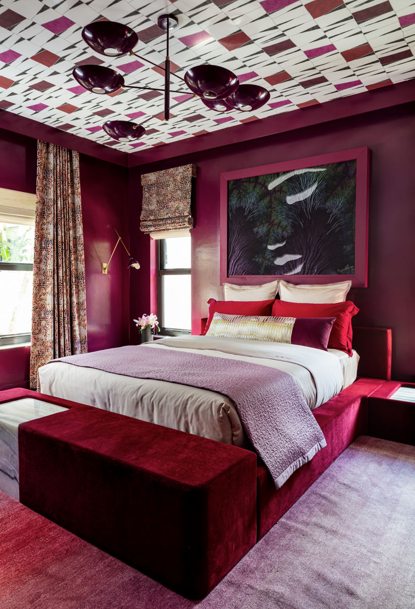 A bedroom in Curated Maximalist style, with burgundy walls, red platform bed with white cover and purple throw, chandelier, and red patterned wallpaper on the ceiling.