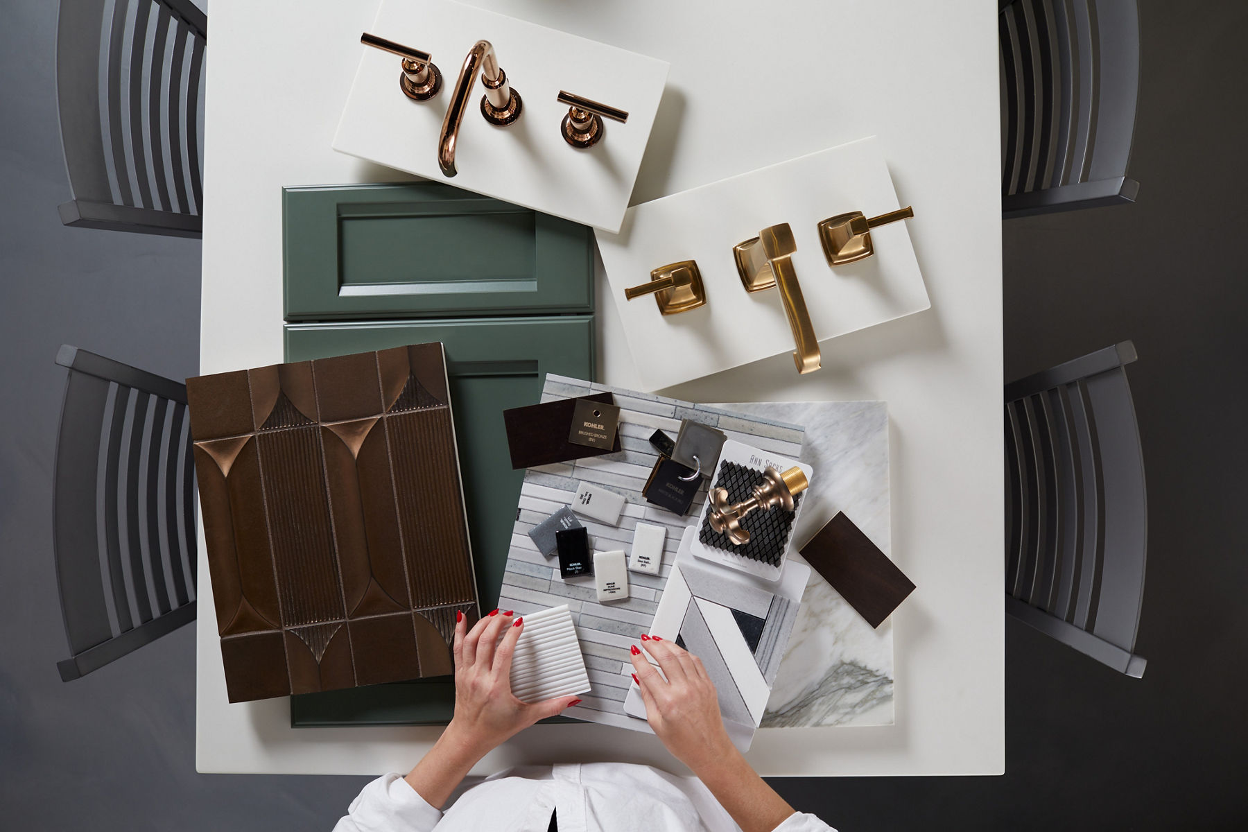Design planning layout featuring brass faucets, cabinet samples, tile swatches, fabric options, and marble slabs, with a person’s hands arranging textured samples on a white surface.