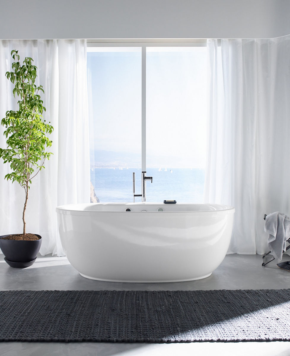 A potted tree stands next to a white KOHLER Sunstruck oval BubbleMassage Air bath with Composed freestanding bath filler and handshower.