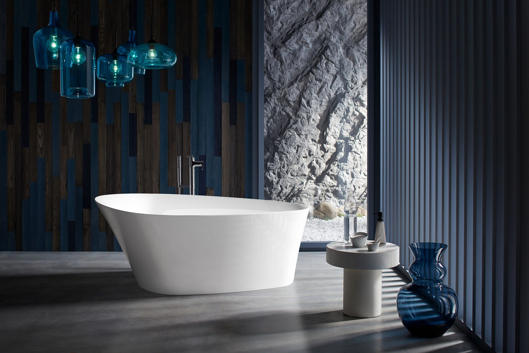 Les lampes suspendues avec abat-jour en verre bleu sont accrochées au-dessus d’une baignoire autoportante blanche Veil de KOHLER.