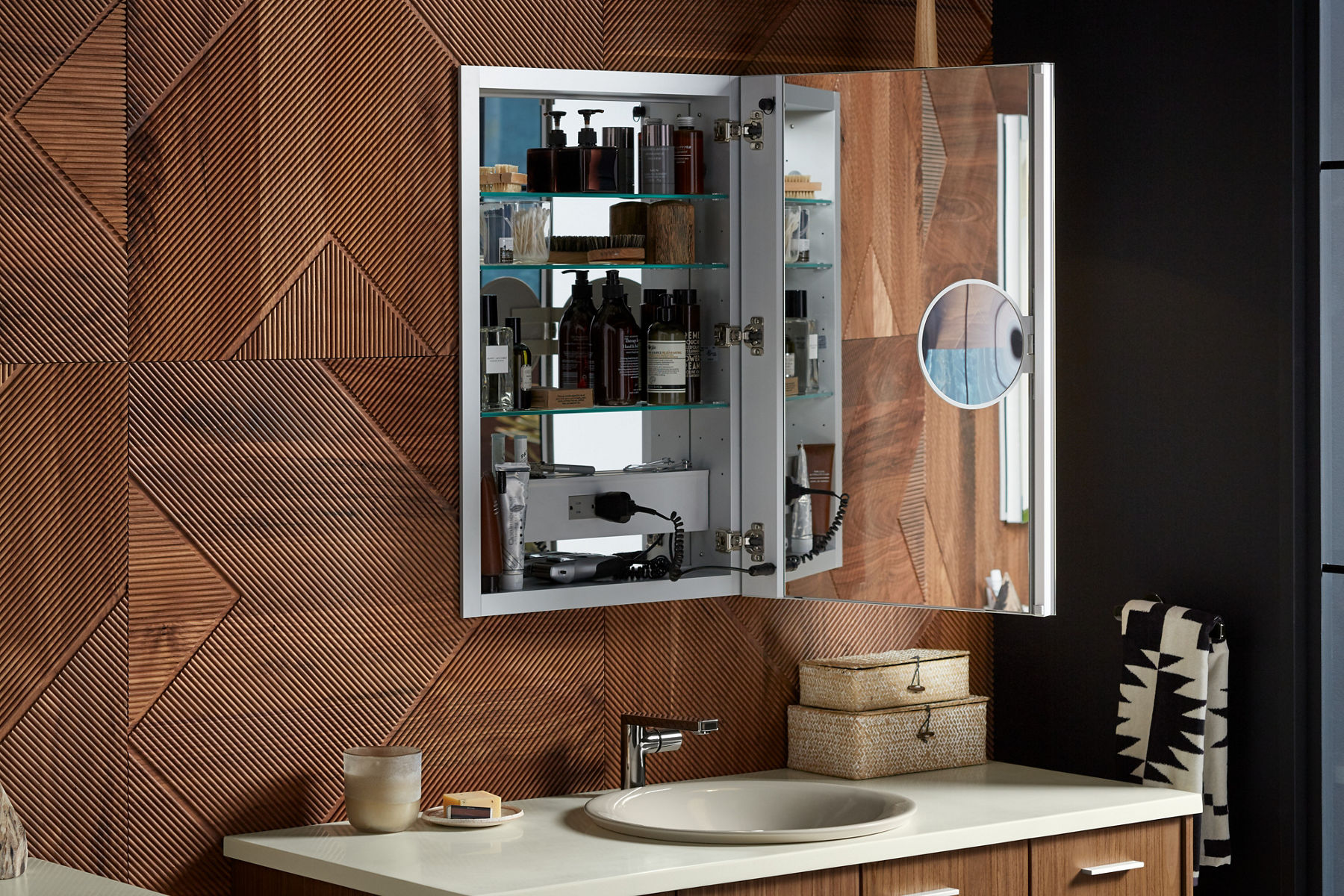 Une armoire à pharmacie KOHLER ouverte avec un intérieur à miroir et un miroir grossissant réglable est suspendue au-dessus d’un meuble-lavabo en bois à dessus blanc avec lavabo encastré.