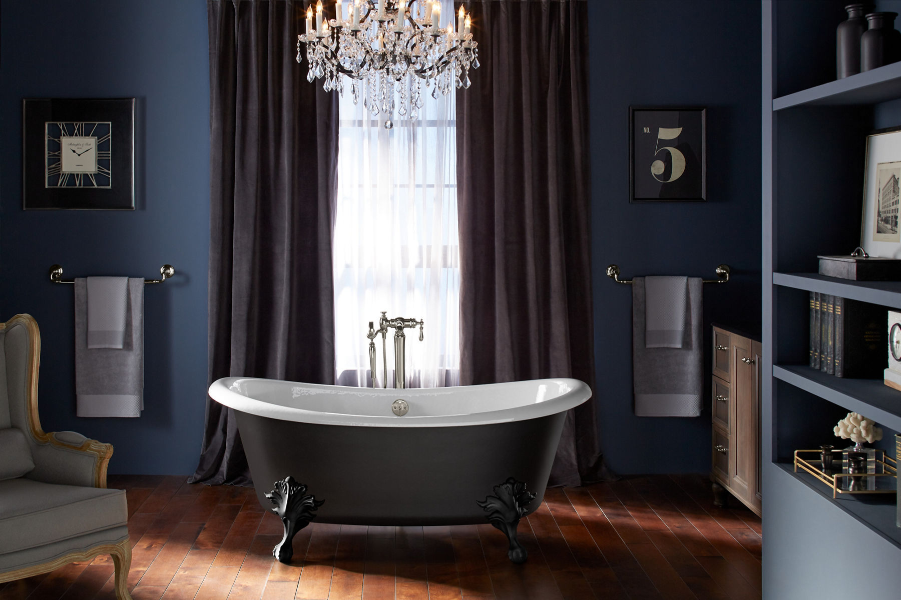 A KOHLER Artifacts Freestanding bath with Iron Black painted exterior and claw feet is the focal point in a dark, dramatic bathroom with a chandelier, floor to ceiling drapes, and towels on holders.