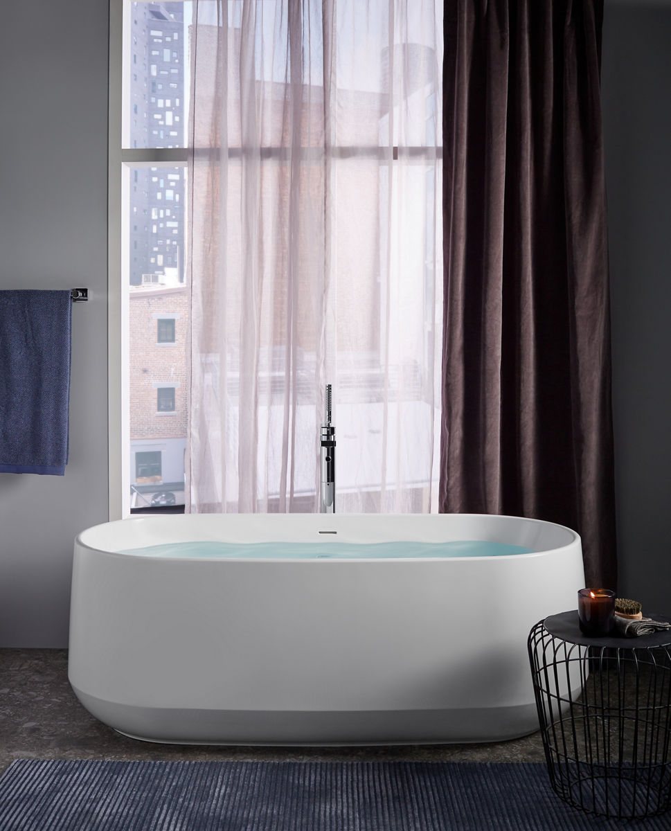 A white KOHLER Ceric freestanding bath and Parallel freestanding bath filler stands before a window with floor to ceiling drapes and city views.