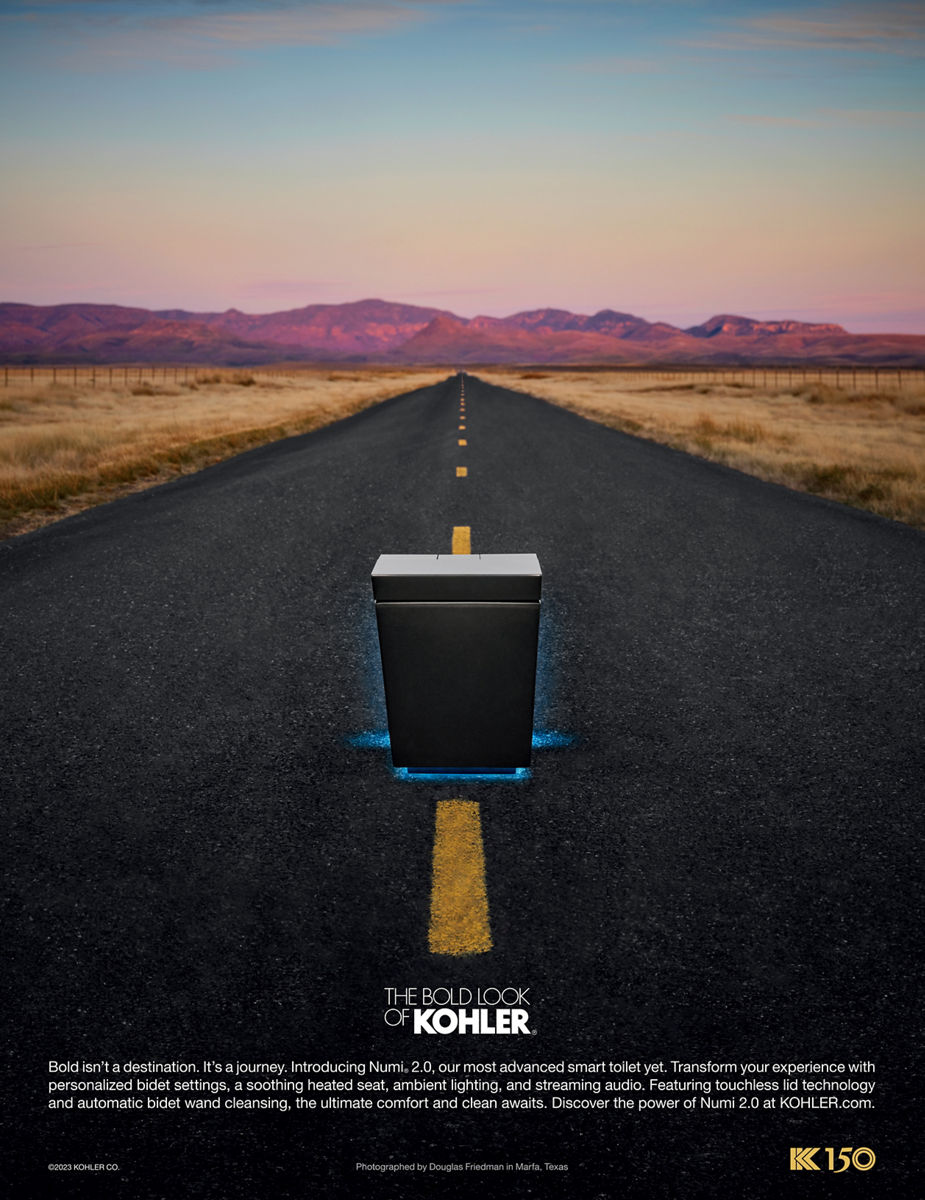 Une publicité imprimée d’une toilette Numi illuminée au milieu d’une route avec des montagnes violettes éclairées au coucher du soleil au loin.