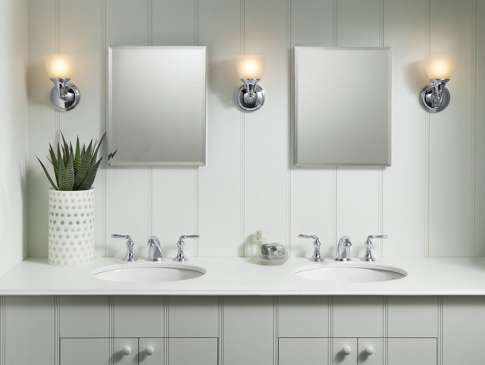 A light gray double vanity against a light gray wall with a white counter top, chrome faucets, mirrors and lighting.